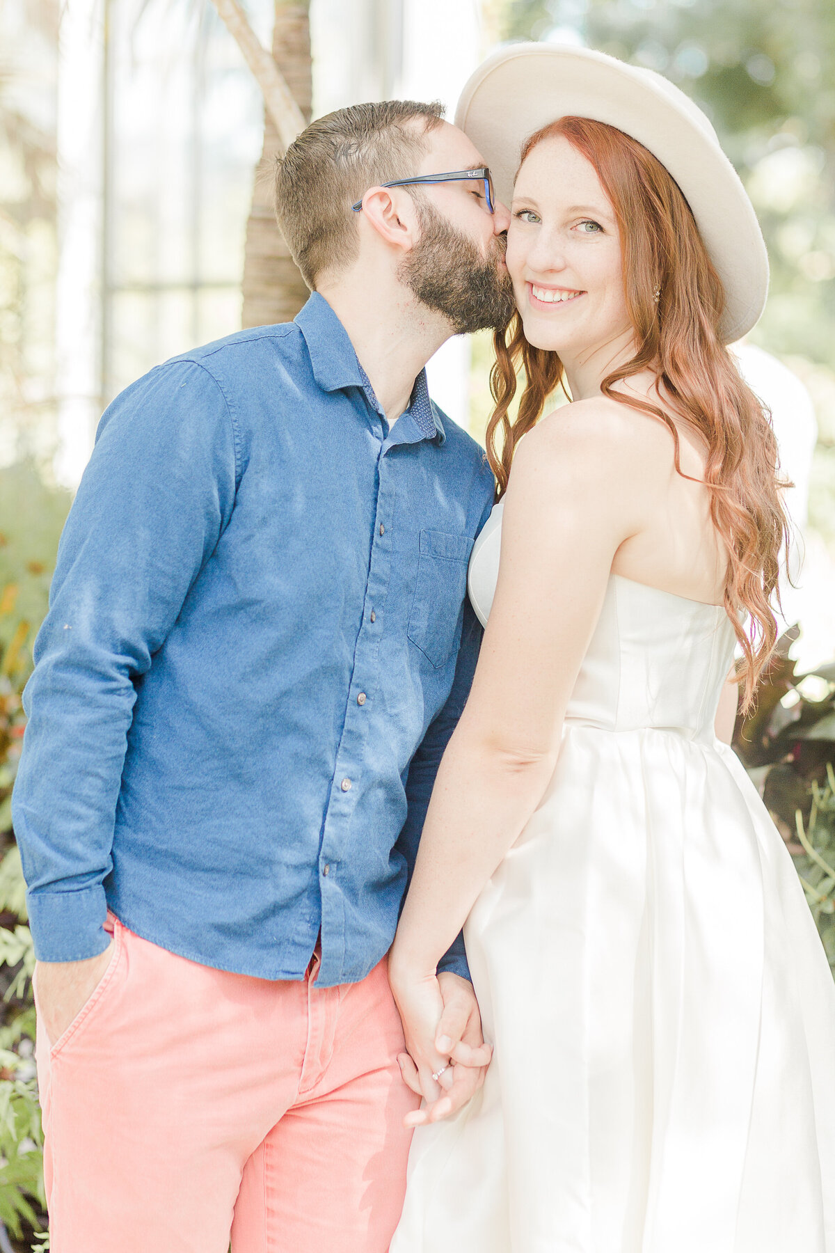 A future bride stands with her back angled to the camera and her fiance facing the camera. Her chest is by his shoulder and they are holding hands. She is looking over her shoulder and smiling at the camera and the soon-to-be groom is kissing her on the cheek. Captured by best RI wedding photographer Lia Rose Weddings