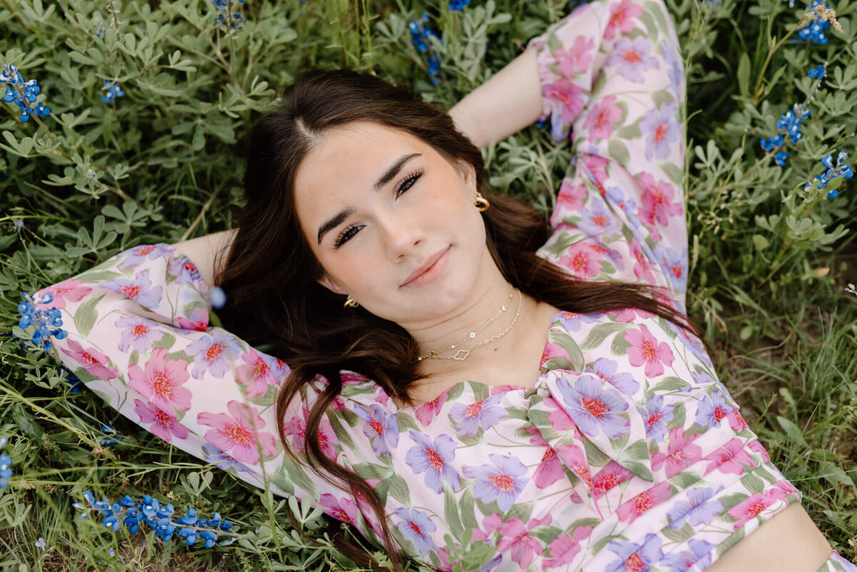 graduate portrait of girl in floral dress lying in a field of East Texas bluebonnets