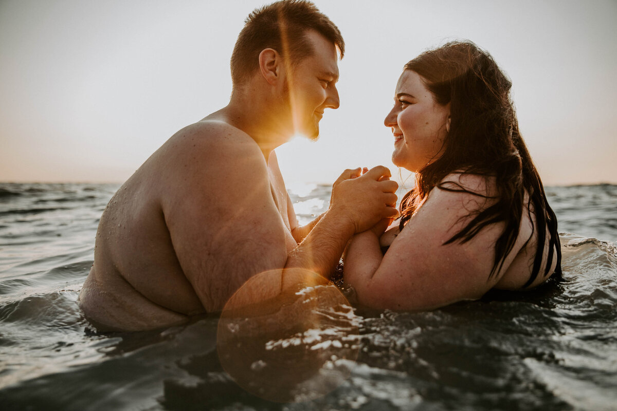 Man and woman sitting in Lake Huron for engagement photos. Lower half of their bodies are in the water, only see their chests. Bride is wearing a black bathing suit. The are facing each other, smiling, and holding hands. The sun is setting behind them creating warm light and sun flare.