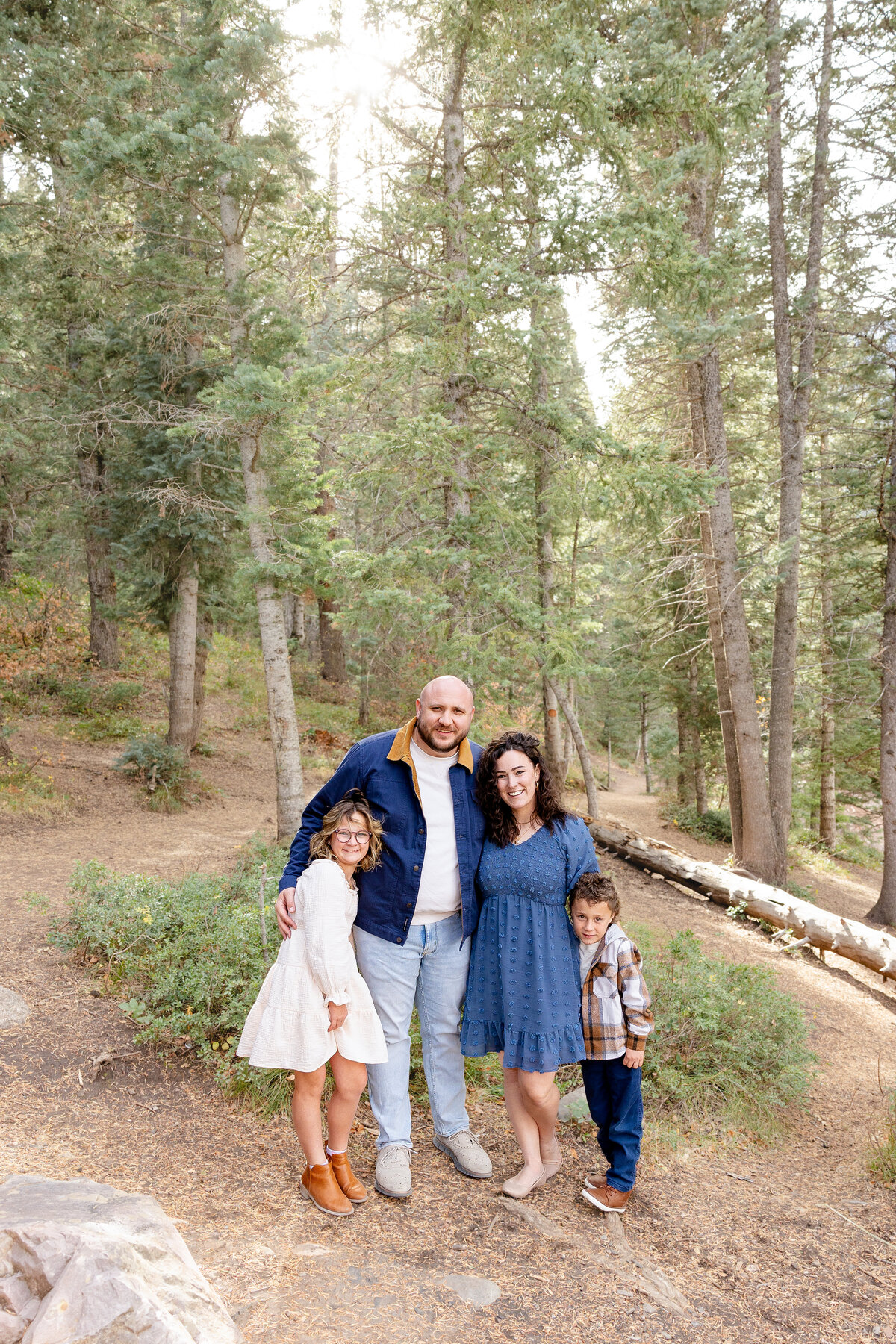 A family of four stands together in a serene forest at Tibble Fork Reservoir, Utah. They are dressed in coordinated blue and neutral tones, smiling warmly at the camera.