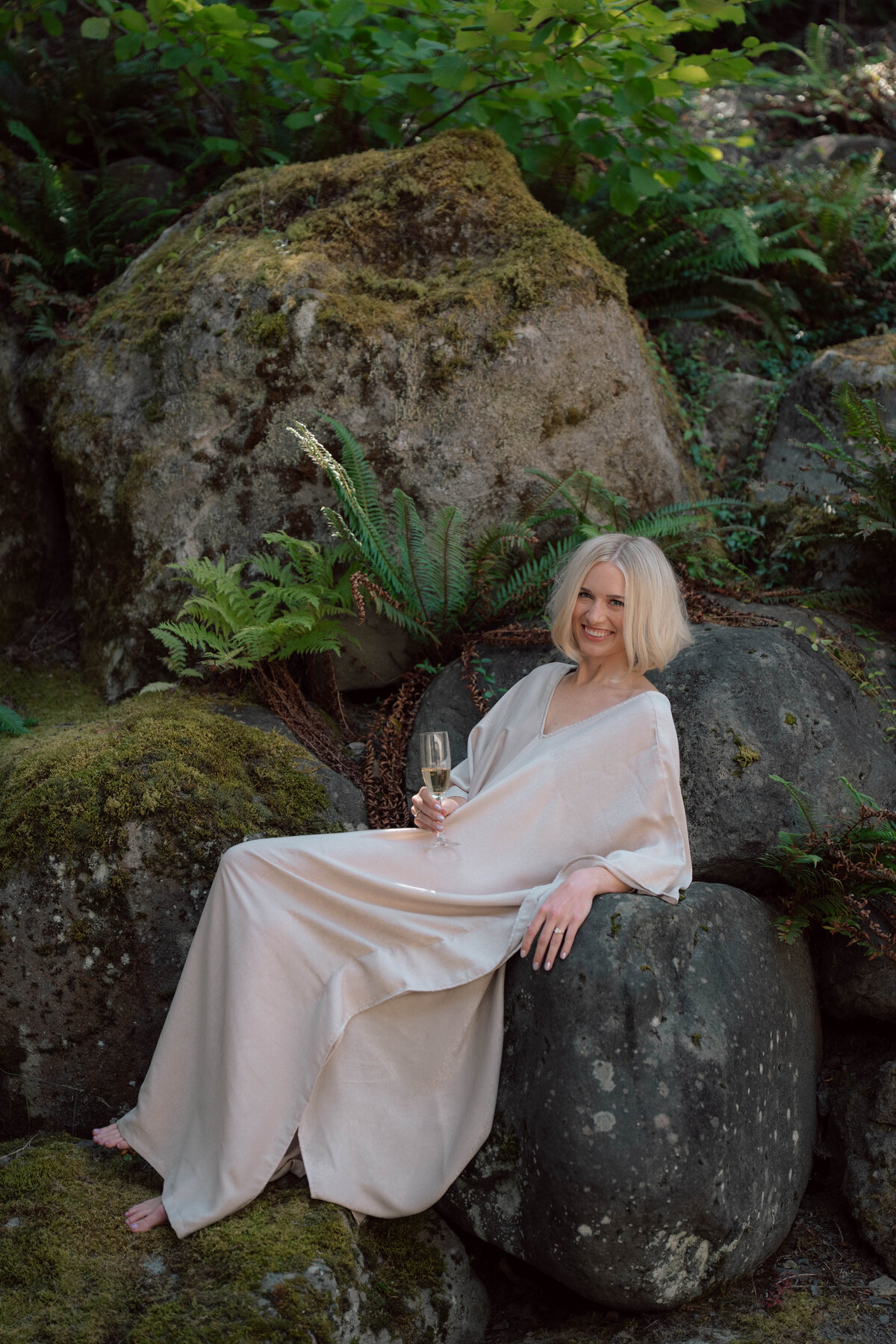 Bride in satin robe holding champagne seated on mossy rocks outdoors