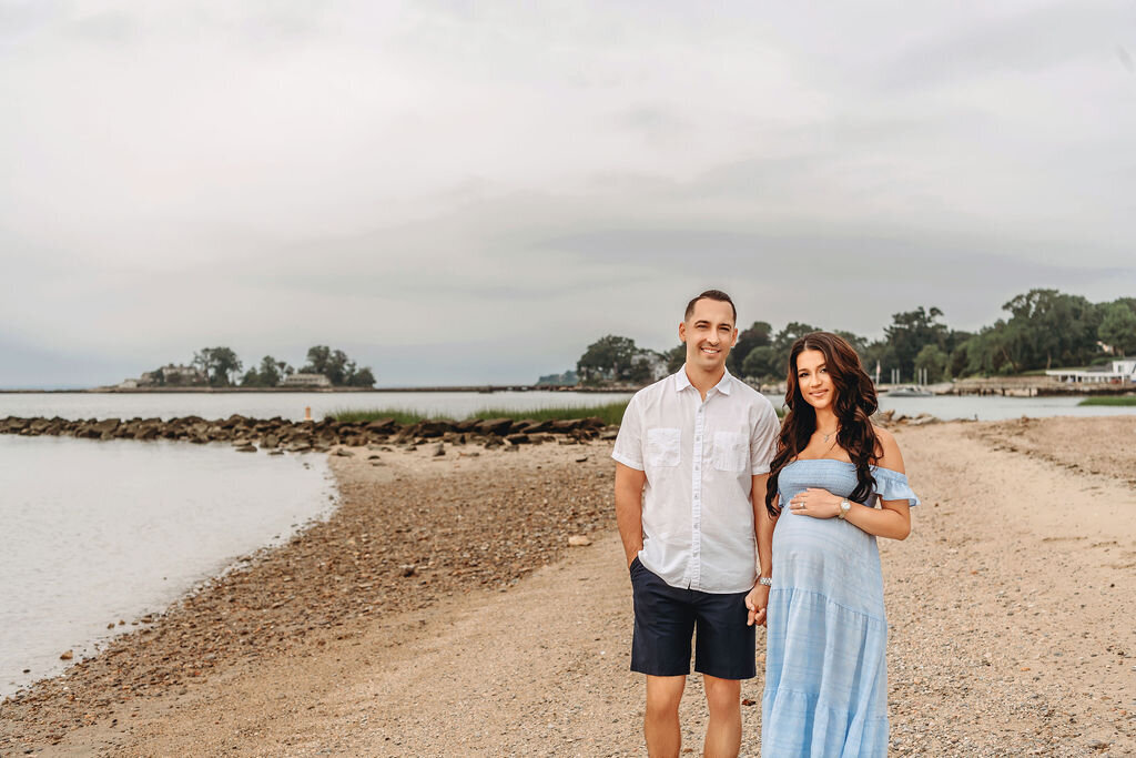 Connecticut maternity photographer photographs couple standing on beach with water and trees backdrop