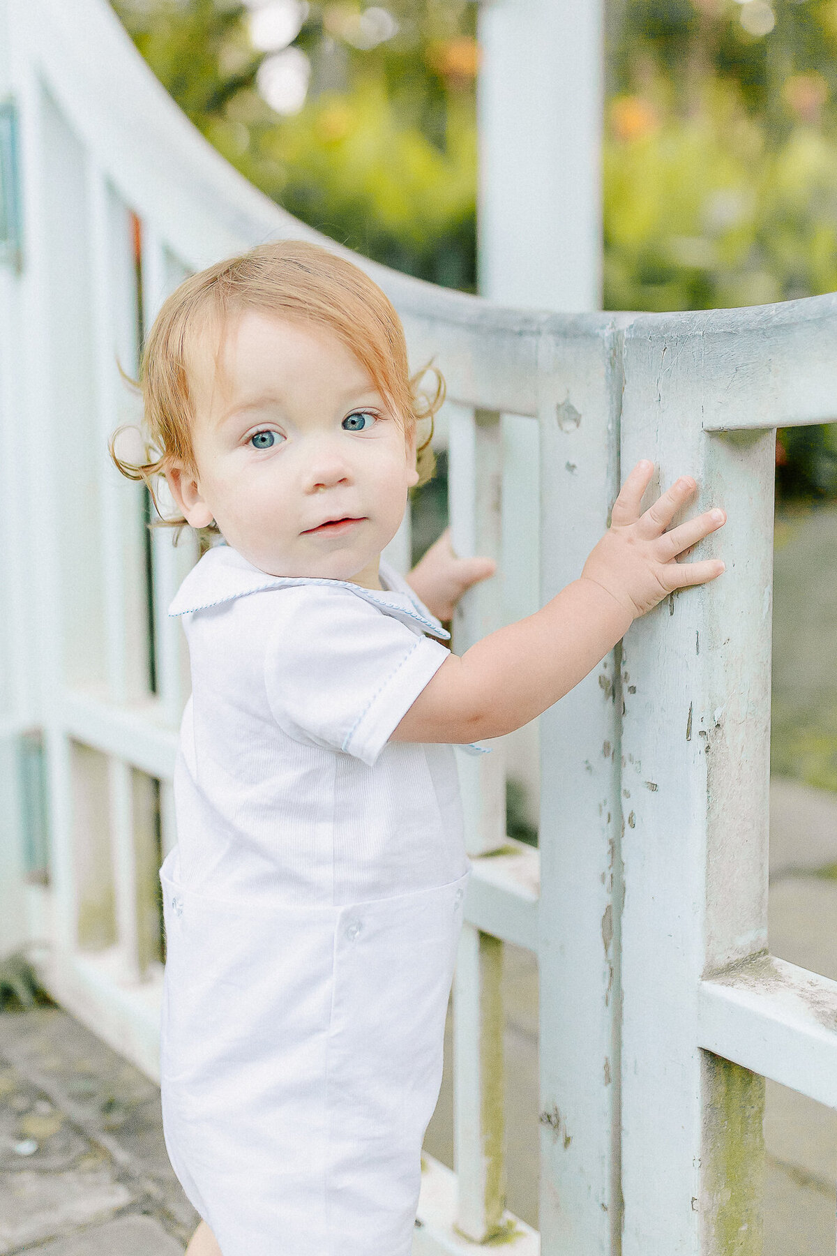 Timeless childhood portrait of a toddler in a classic white outfit, holding onto a garden gate with curiosity and wonder—captured by a Birmingham, AL family photographer