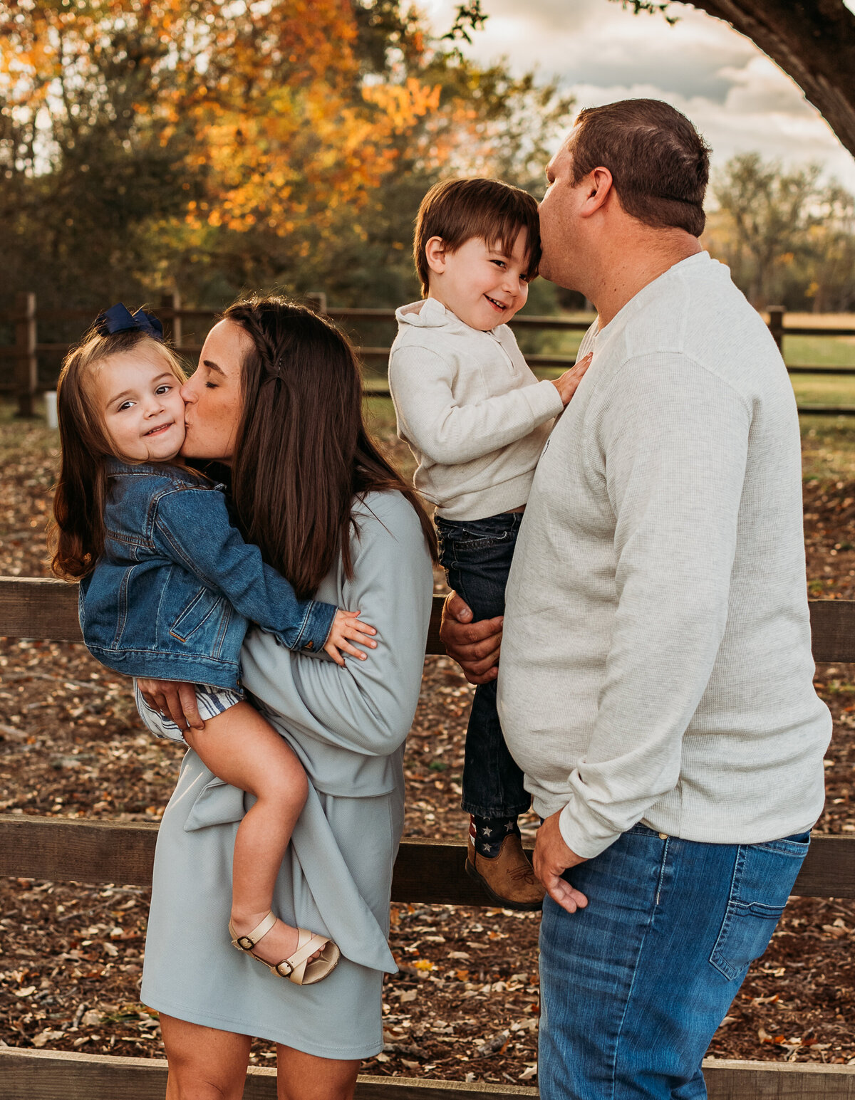 A mom and dad kiss their young children with fall foliage in the background.