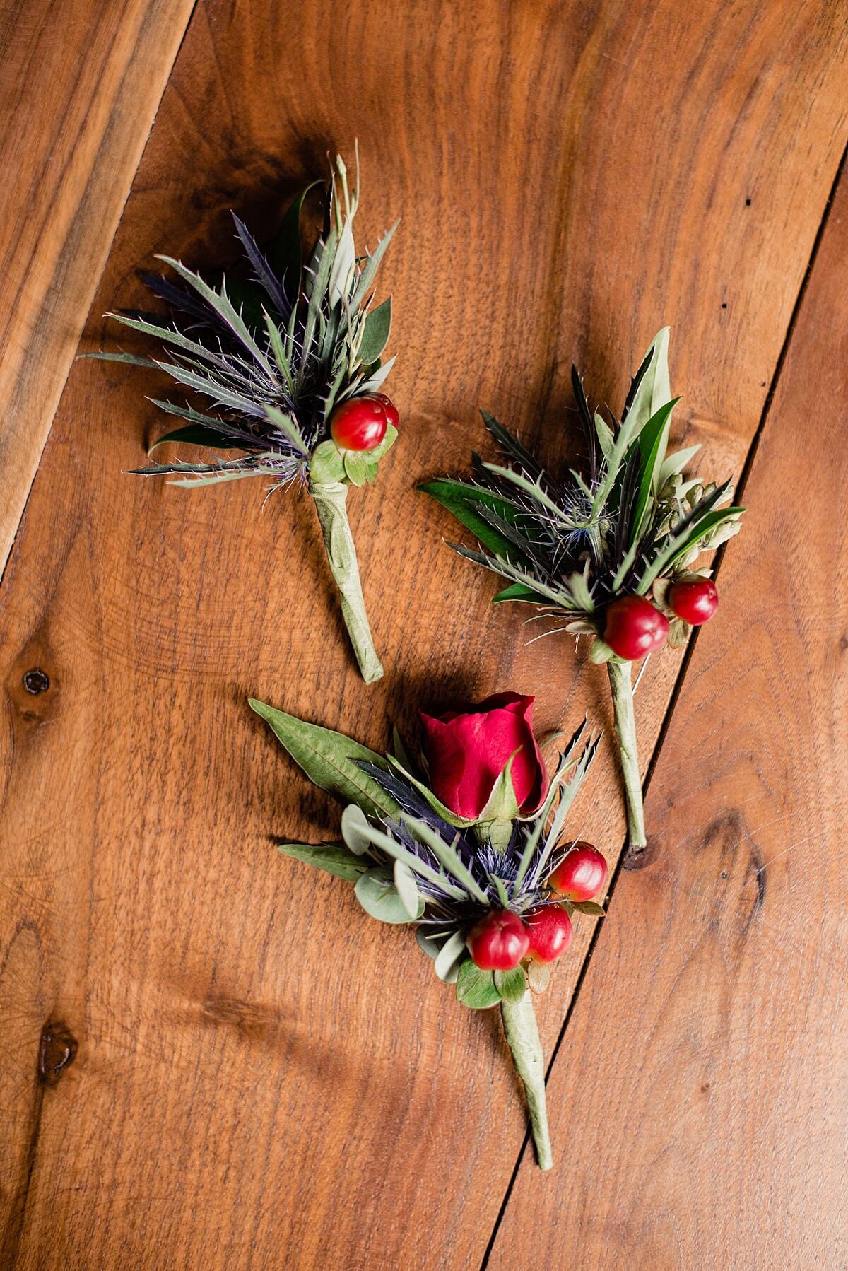 Boutonnieres with red berries, blue thistle and a red rose sit on a farmhouse table.
