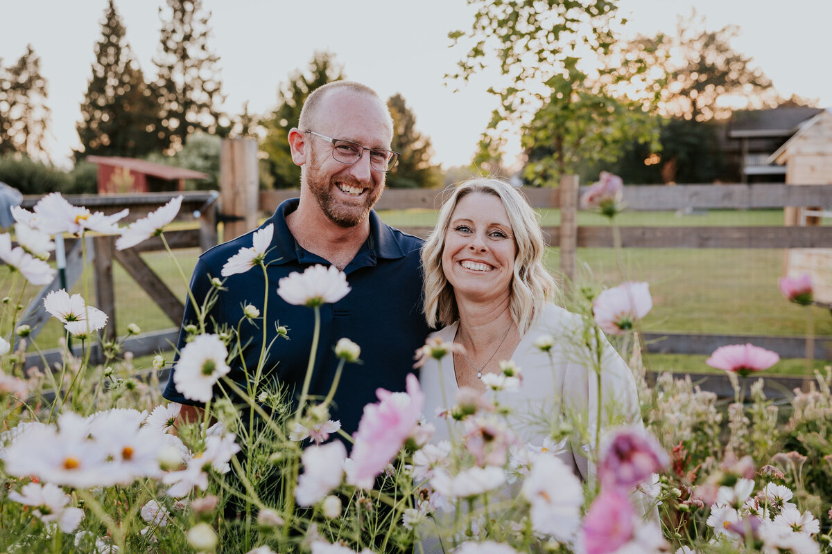 Couple smiles for the camera while standing behind row of tall purple and while flowers.