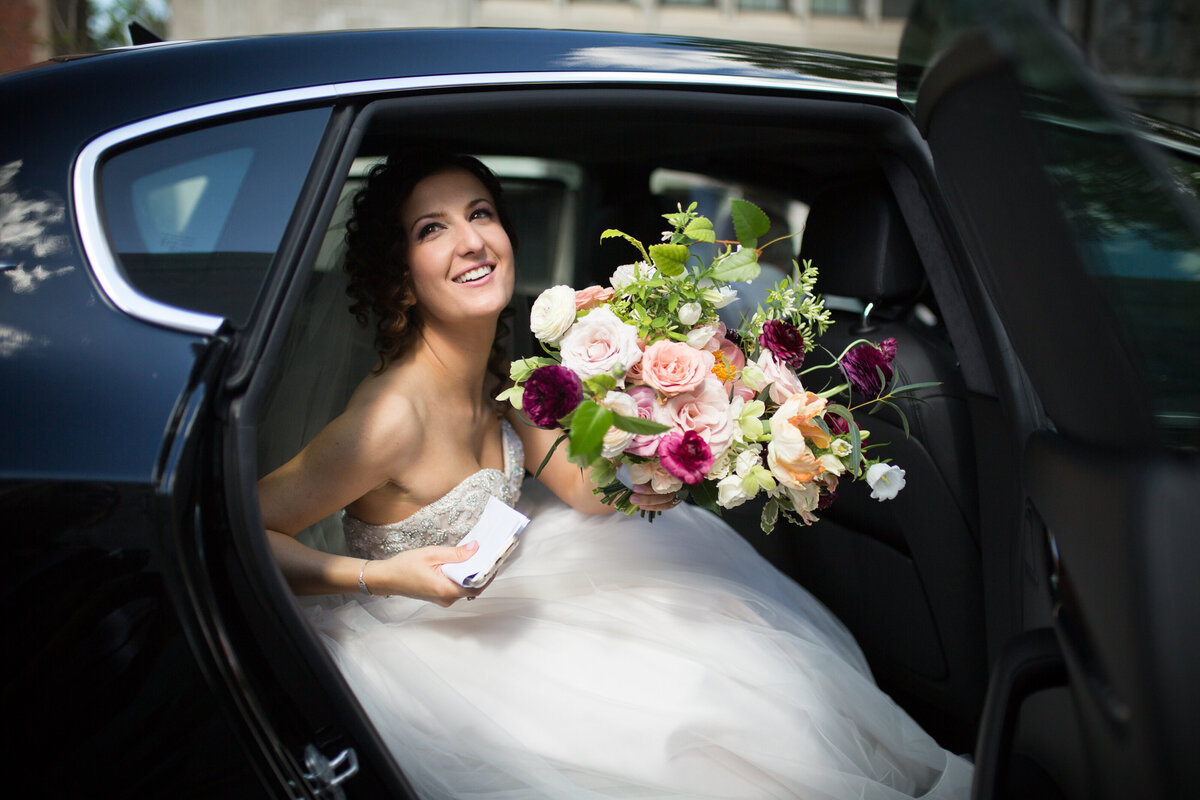 A bride smiles exiting a limousine holding a large, bright bouquet of flowers.