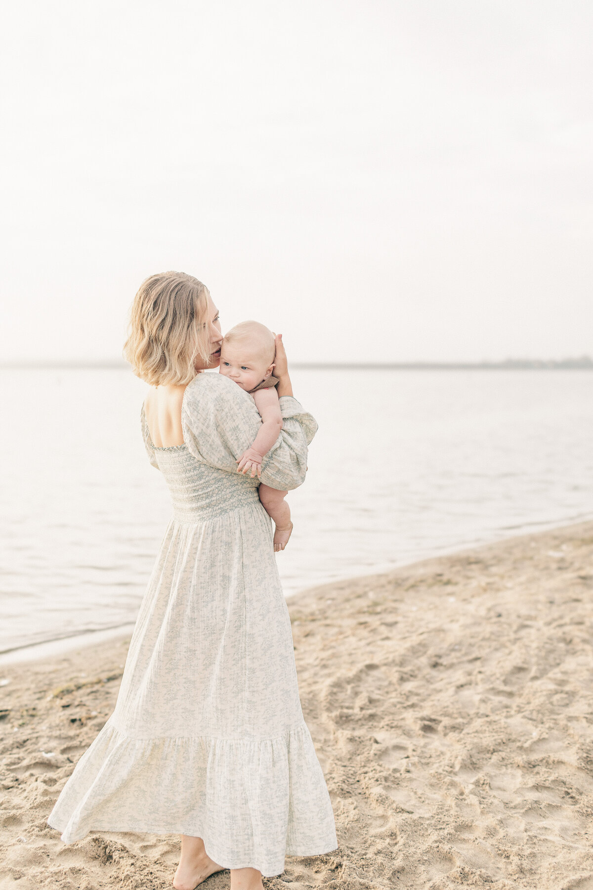 Mother holding her baby boy with her back to the camera soothes him with a lullaby as they look out over the water during their Kansas City beach session.