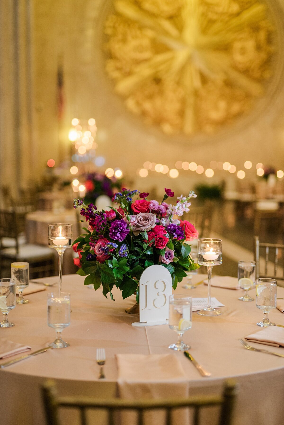 A detail shot of a reception table at a wedding reception at the Hall of State at Fair Park in Dallas, Texas. The large, round table is covered by a tablecloth and has multiple napkins, set of cutlery, glasses around the edge. In the middle of the table is a large bouquet of colorful flowers and a table number sign, and candles. Other similar tables can be seen in the background and a large gold crest can be seen up on the background wall.