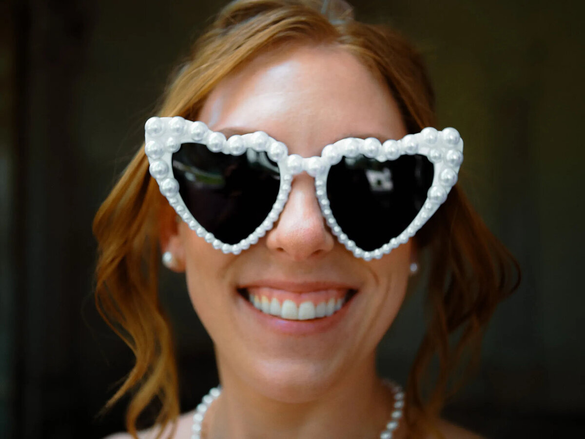A smiling woman with wavy reddish-brown hair wears large, heart-shaped sunglasses decorated with pearls. Captured by a talented NJ wedding photographer, she also wears pearl earrings and a pearl necklace, standing against a blurred background.