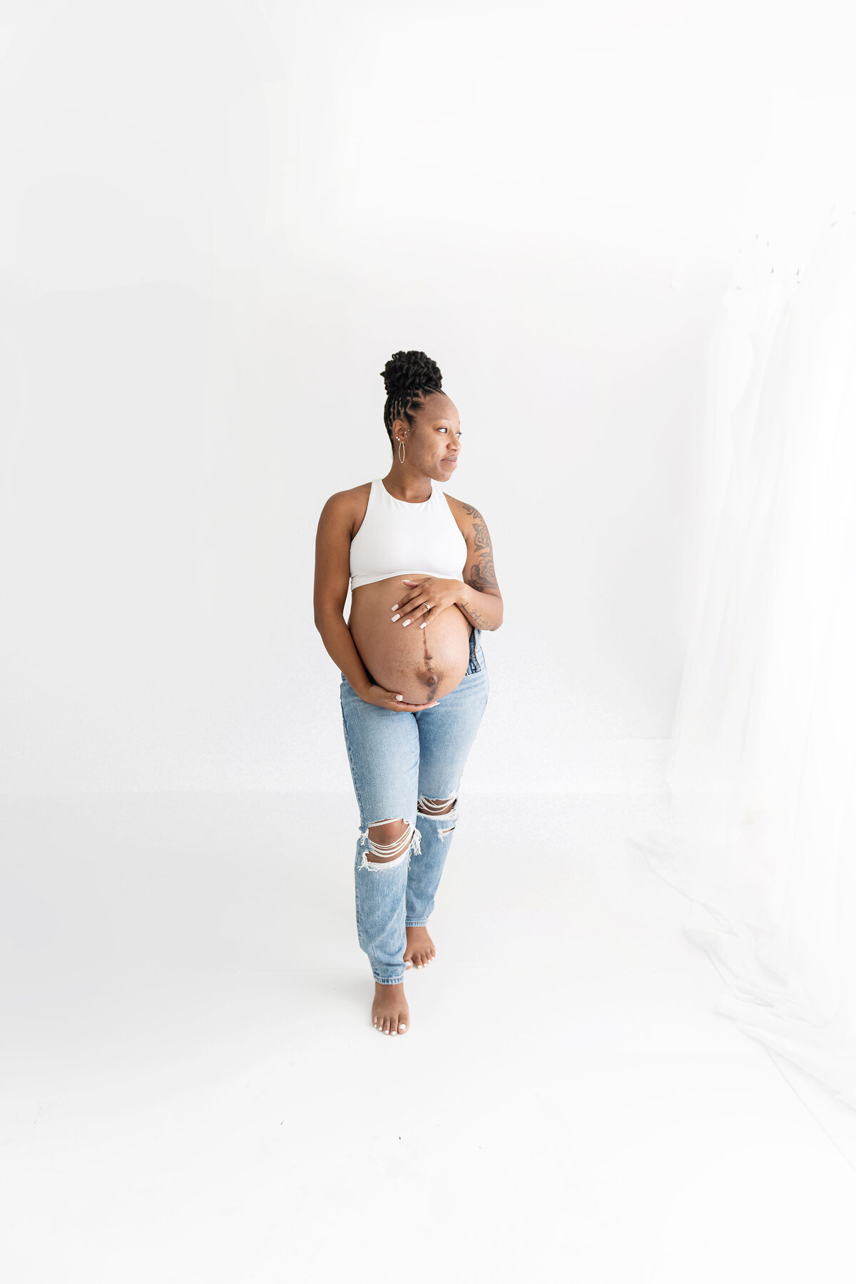 A mother to be stands in a studio holding her exposed bump in jeans and a white top