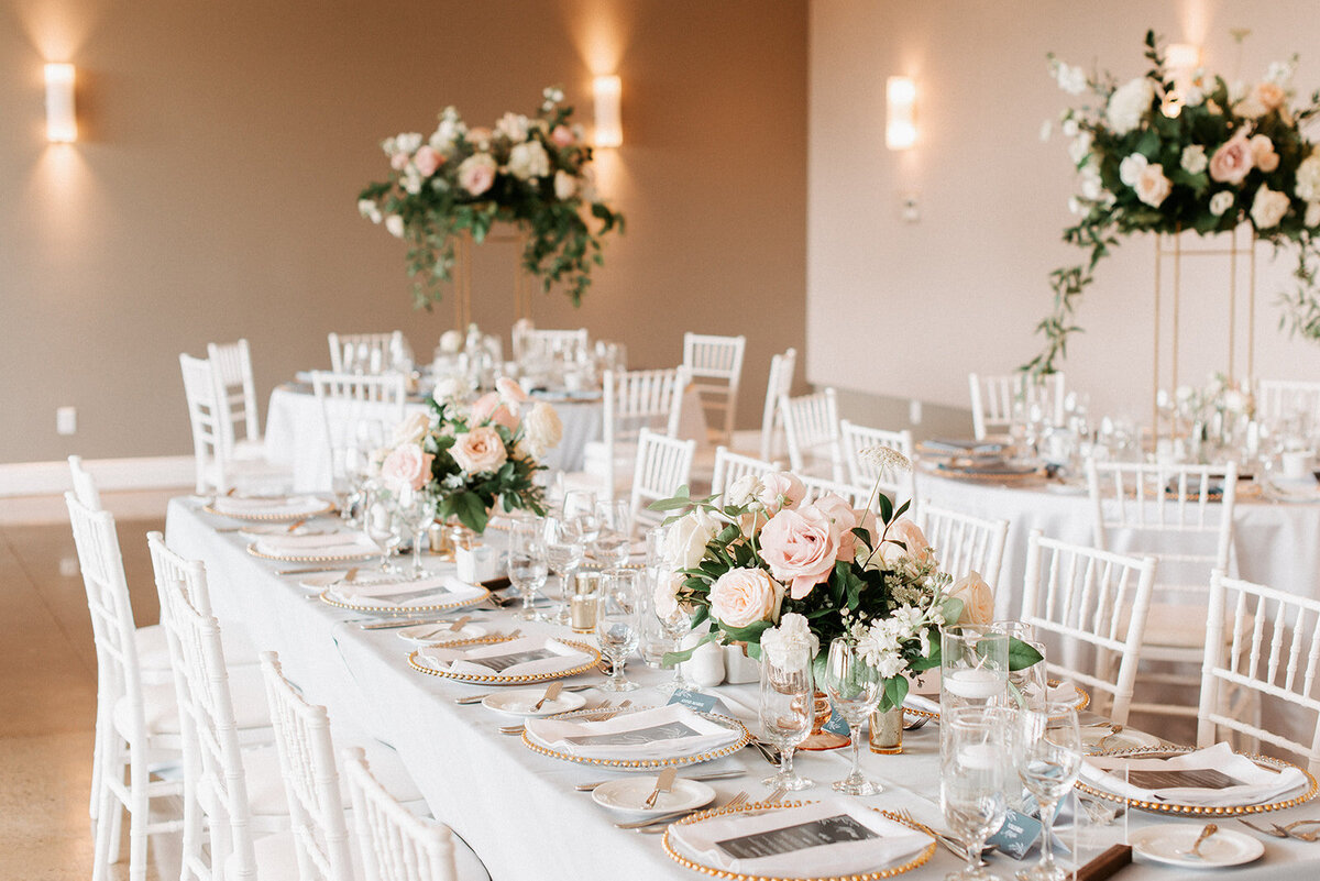 A long guest table with low blush floral centrepieces and dusty blue linens sits in the middle of Le Belvedere's room with tall centrepieces in the background for a wedding  in Wakefield Quebec.