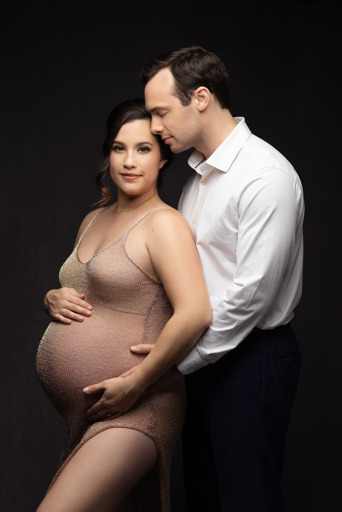 Lovely couple standing together, the man kissing the top of the woman's head and holding her belly. The woman wearing a sheer blush gown looking towards the camera with a soft smile on her face. Captured by Dallas maternity photographer Lindsay Walden.