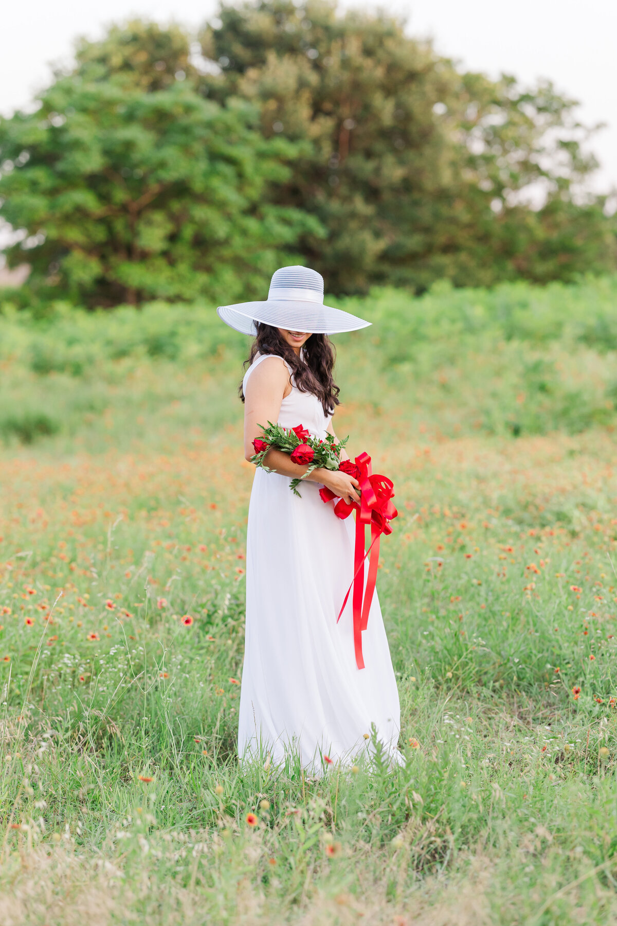 Ursuline Academy graduate in a flowing white dress and wide-brimmed hat stands in a wildflower field holding a bouquet of red roses tied with a red ribbon. A serene and elegant portrait set in nature.