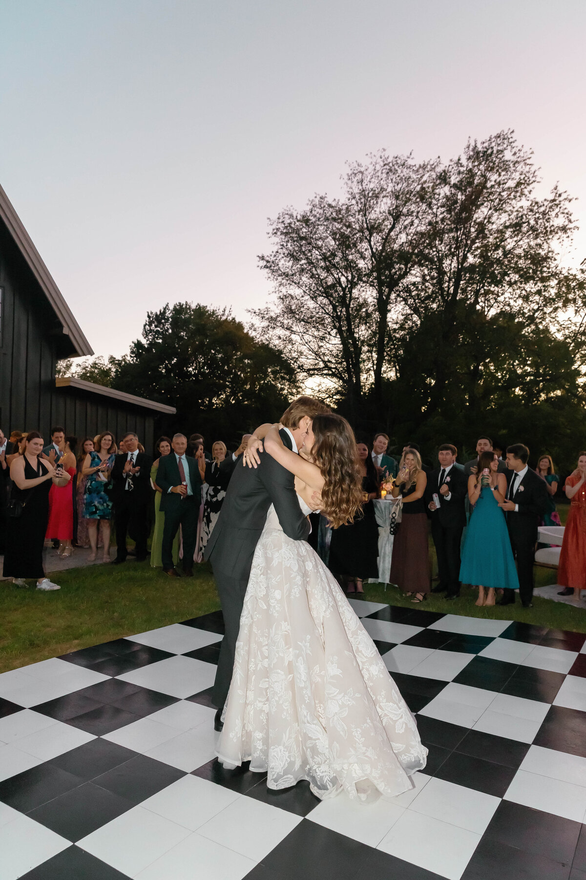 Bride and groom sharing their first dance on a black-and-white checkered dance floor at their outdoor Arkansas wedding reception, surrounded by cheering guests at dusk. The bride wears a lace ballgown, and the groom wears a classic black suit as they embrace beneath soft evening light.