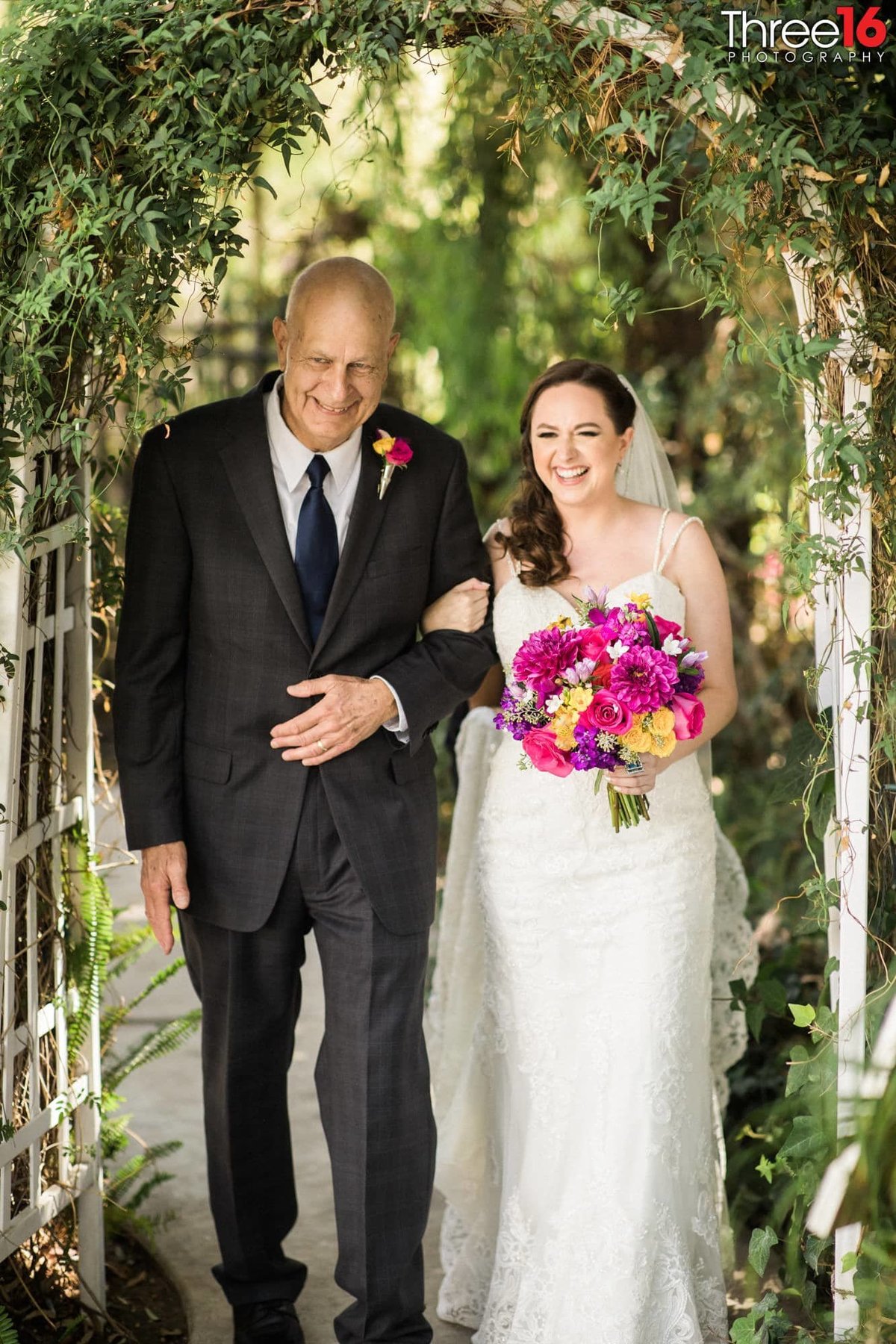 Father of the Bride escorts under an floral archway heading towards the altar