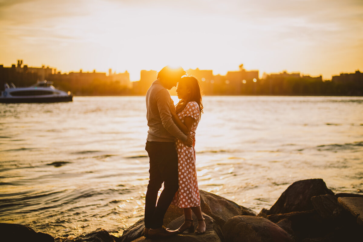 An engaged couple embrace on a jetty rocks at domino park in brooklyn at sunset