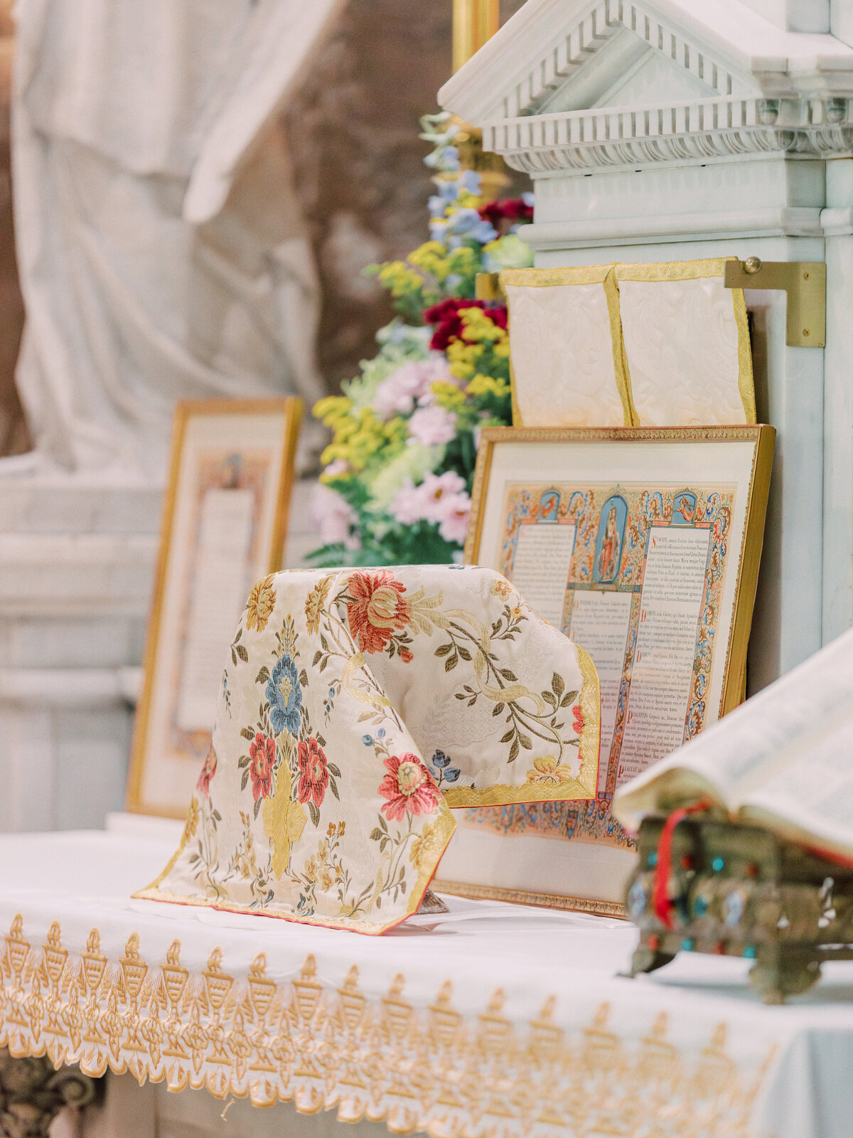 Ornately decorated altar for a traditional Latin Mass, featuring embroidered altar linens, a floral chalice veil, a framed altar card with prayers, an open missal, and colorful floral arrangements in the background at St Leos Oratory in Columbus