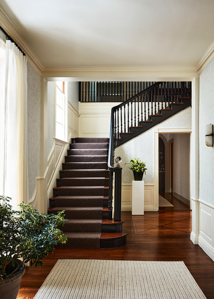Interior decorator in Boston updates a historic Concord home with a bold black staircase and neutral wallpaper, blending classic charm with modern design.