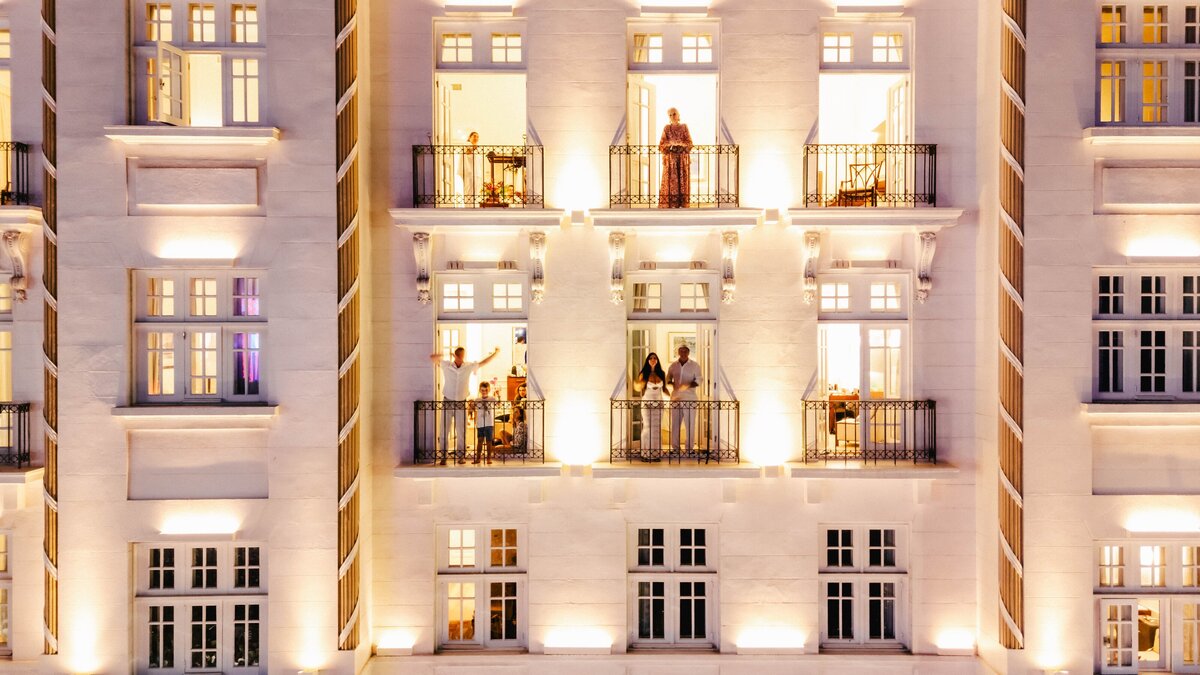 A stunning illuminated boutique hotel with elegantly dressed guests enjoying private balconies at night. Experience bespoke luxury accommodations, high-touch concierge services, and exclusive stays curated by Nadia Forte Travel & Events.