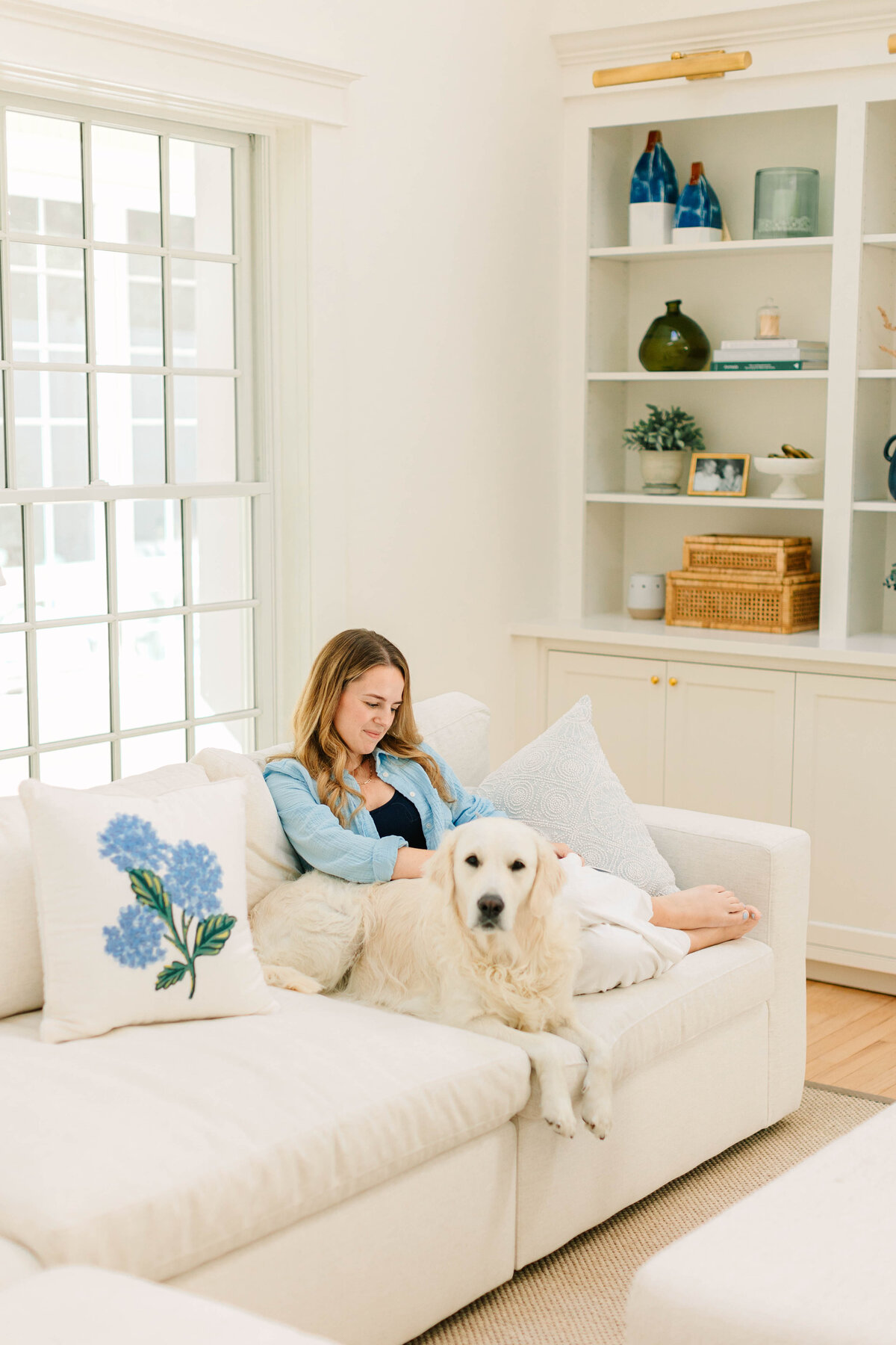 A woman in a light blue shirt and white pants lounges on a cozy white sofa, smiling as she relaxes with her golden retriever. The bright living room features built-in shelves decorated with books, vases, and woven baskets, creating a serene and inviting space.