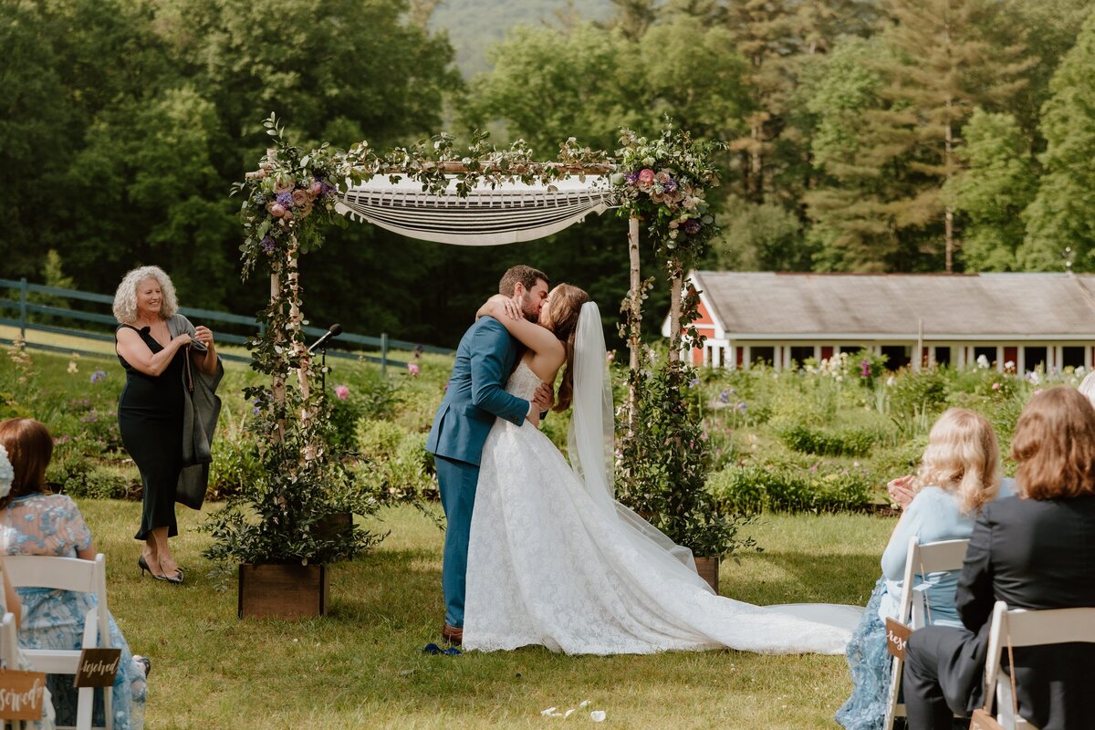 Bride and groom kiss beneath a floral chuppah during a romantic outdoor ceremony surrounded by gardens, mountain views, and applauding guests.