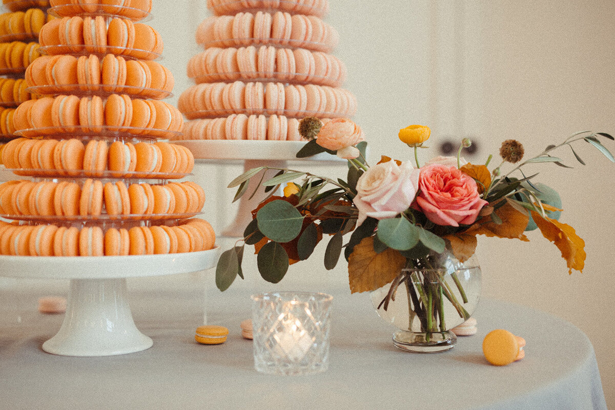 Two cake stands filled with macaroons atop a rounded table with a flower arrangement and a lit candle.