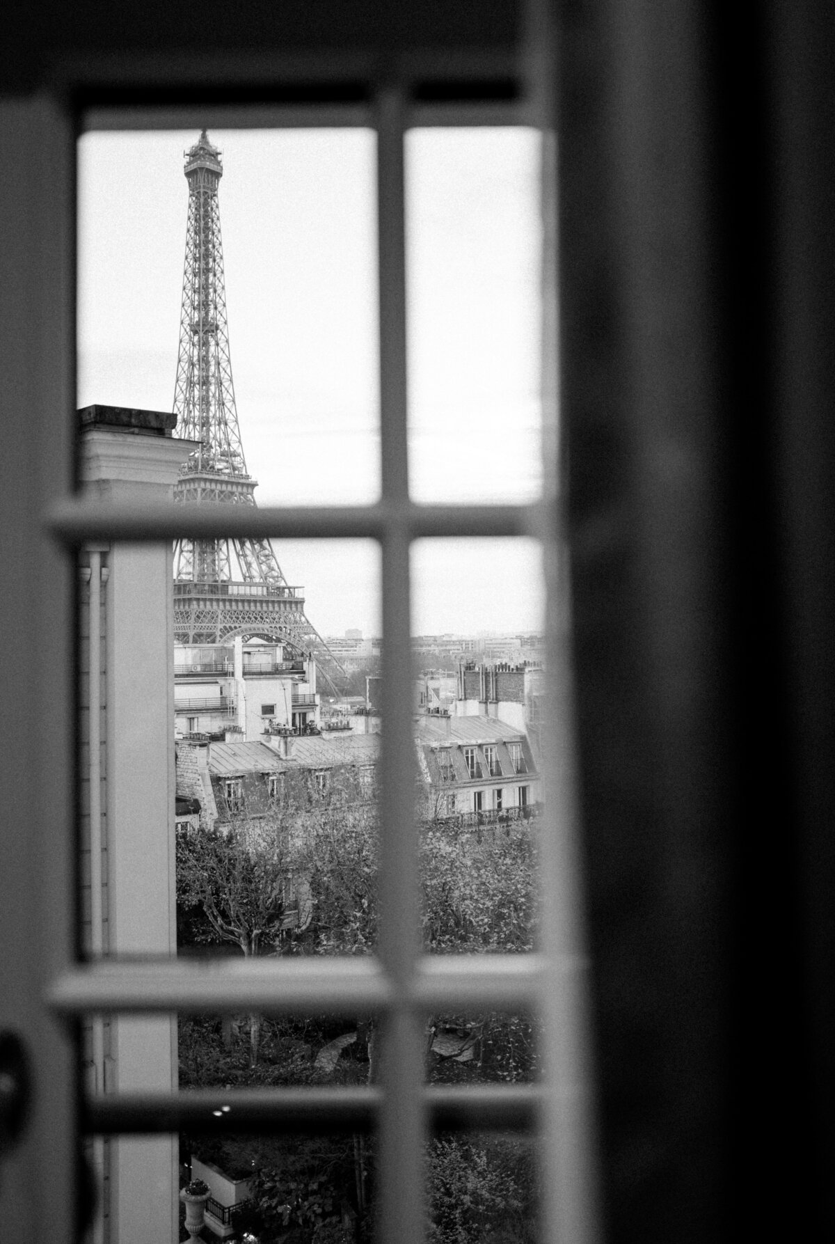 The photo was taken at Shangri La Hotel & Ritz Paris Hotel in Paris, France, by photographer Matteo Coltro. Zen Film Works also contributed to the stunning visuals captured in this image. It is a black and white photograph taken through a window with panes, showcasing the breathtaking view of the Eiffel Tower in the background. The picture elegantly captures the Parisian rooftops and lush trees in the foreground, with the iconic structure majestically rising up behind them, creating a mesmerizing contrast of urban and natural elements.