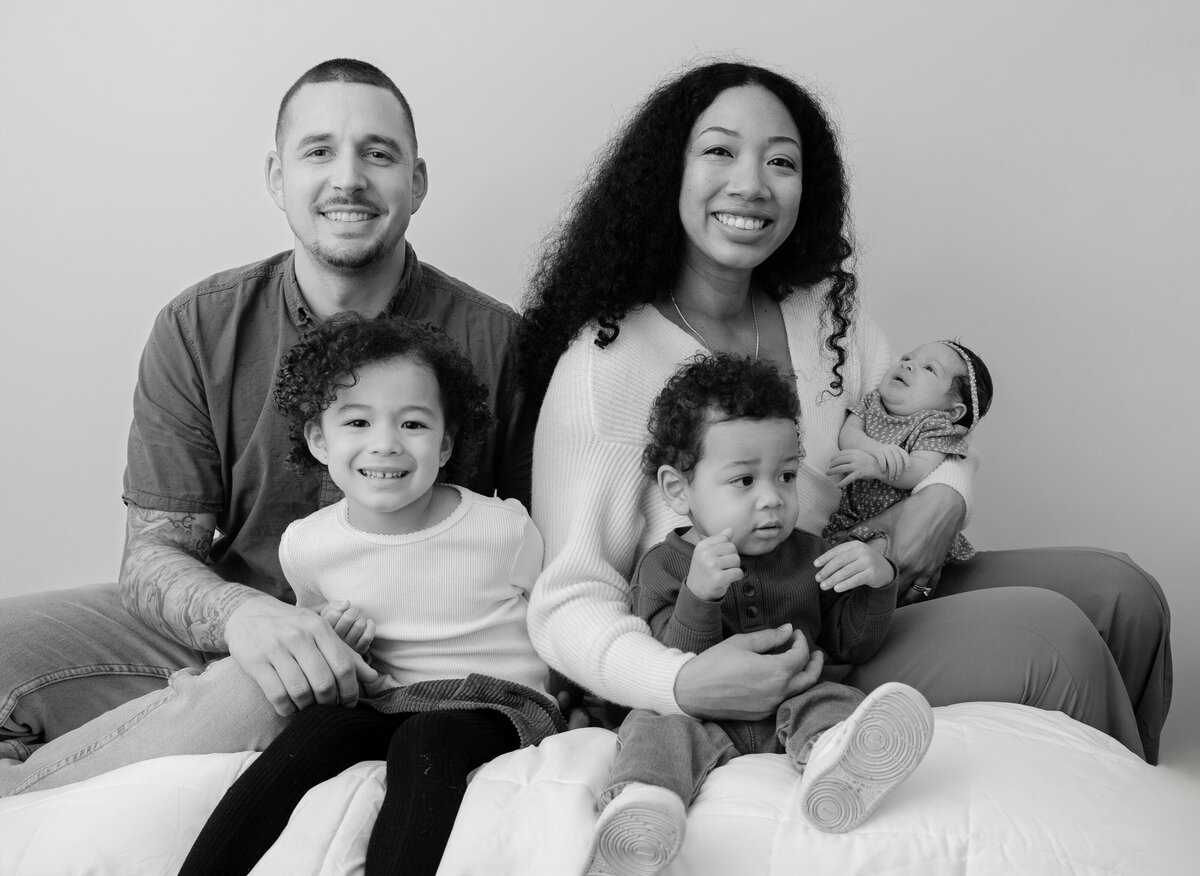 Black and white photo of newborn baby girl held in mom's arms with mom  hugging son and dad sitting down next to mom with daughter in front. 
