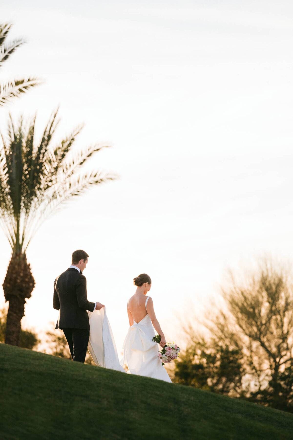 Bride and groom walking together on a sunlit hill surrounded by palm trees, captured by Scottsdale wedding photographers in Arizona.