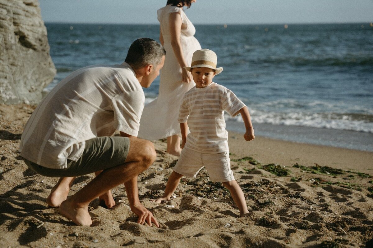Le père accroupi regarde son fils, prêt à jouer. La mère enceinte en robe blanche marche sur la plage. Photo par Kamescop, photographe grossesse.