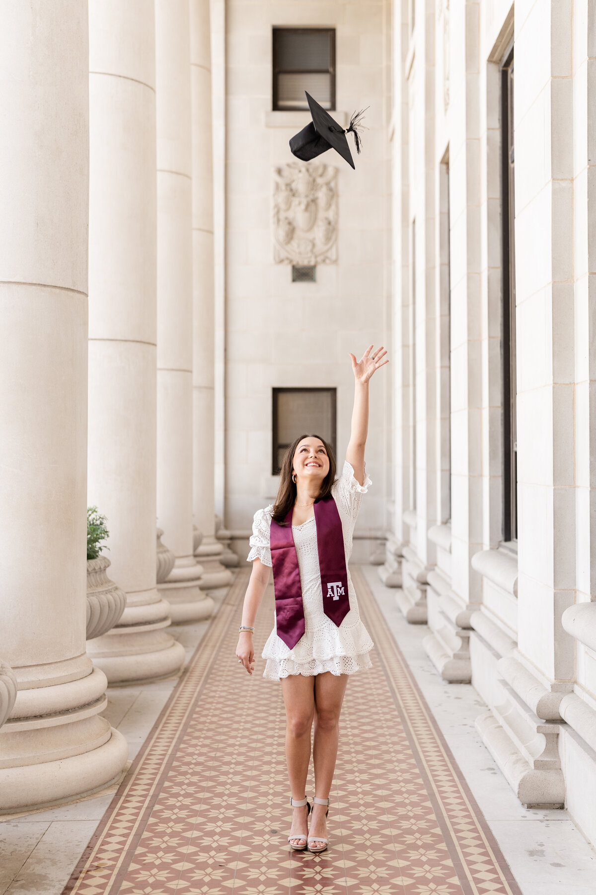Texas A&M senior girl throwing up cap while wearing white dress and maroon stole in the columns of the Administration Building