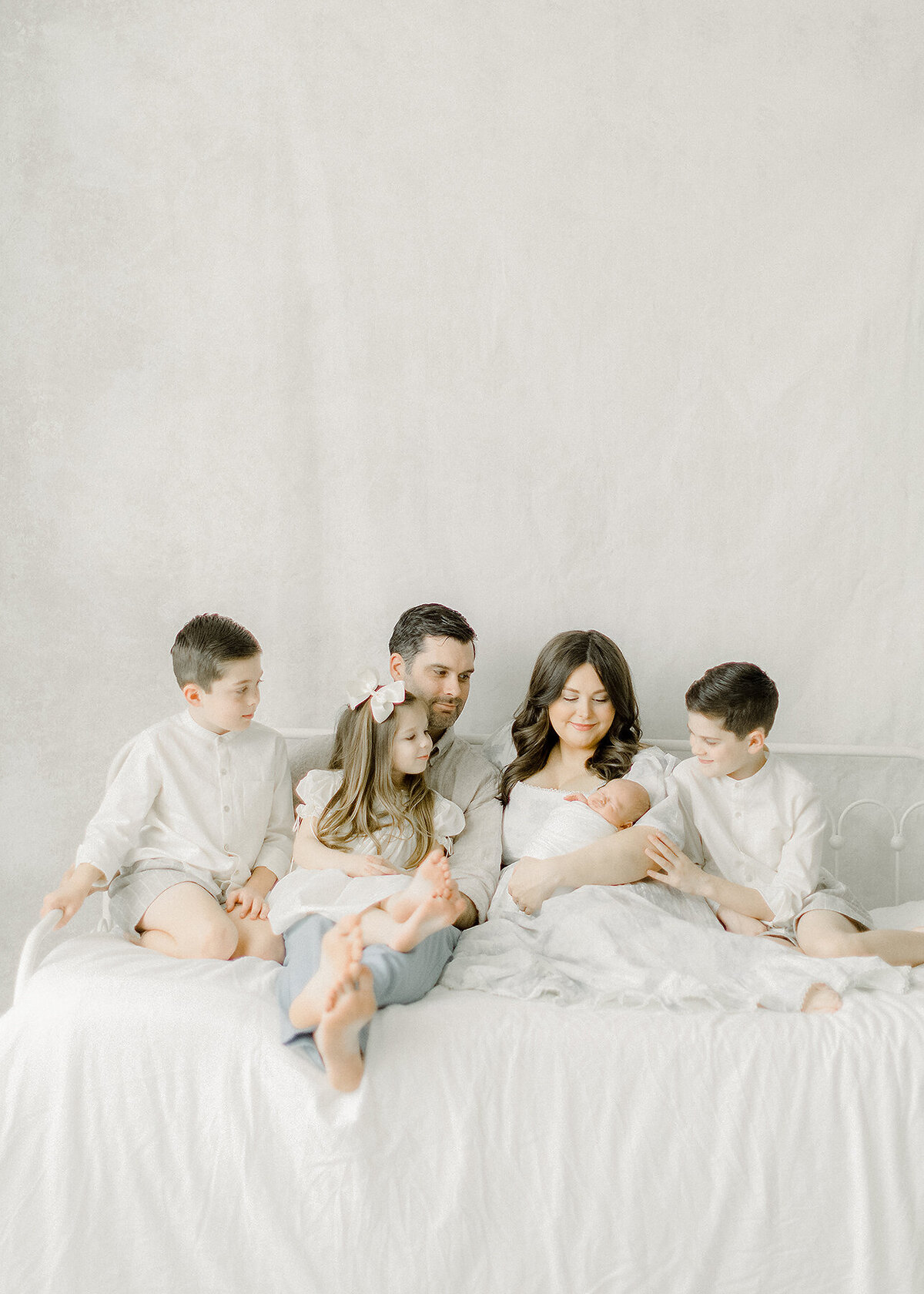 Beautiful Dallas TX family sitting on a bed together in a DFW photography studio while they are leaning in and as the siblings are looking at their newborn baby brother.
