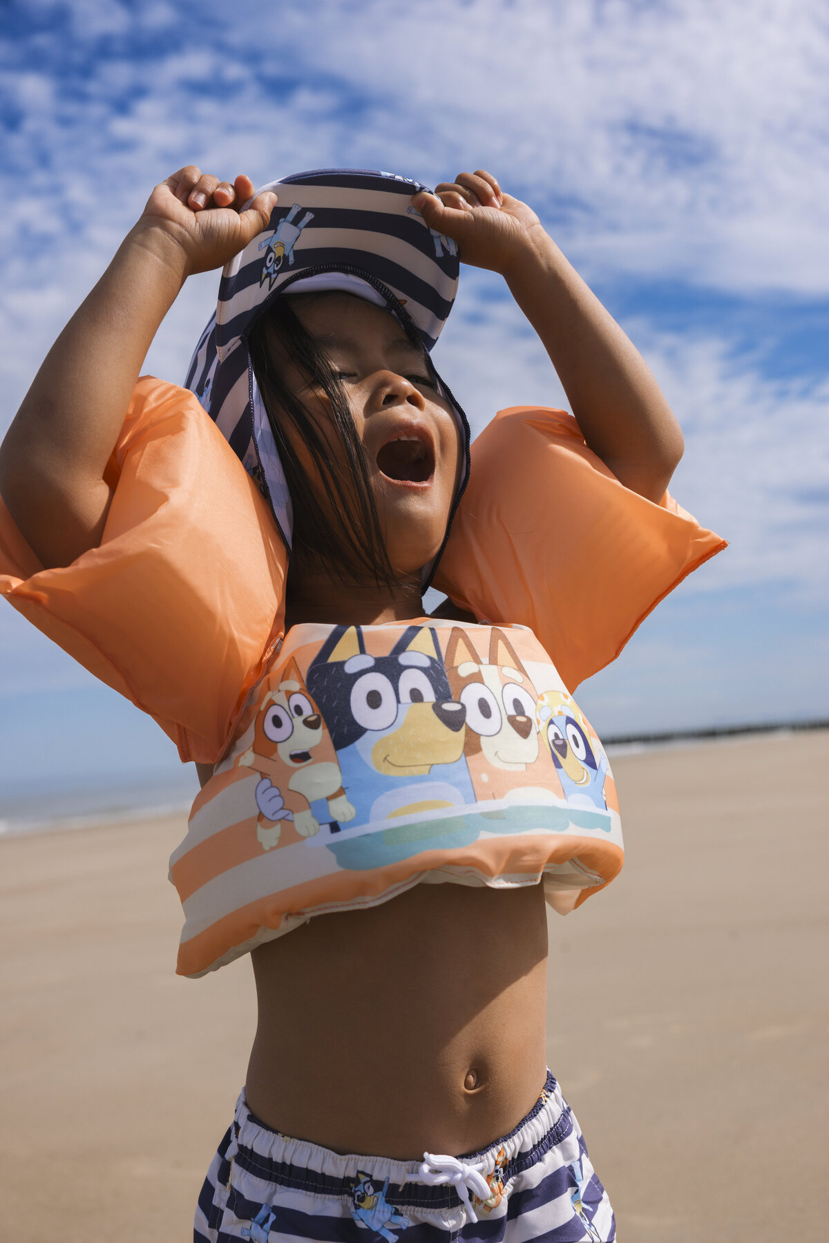 A joyful young girl wearing a Bluey-themed float vest and striped swim shorts from Swim Essentials, raising her arms with excitement on a sunny beach day.