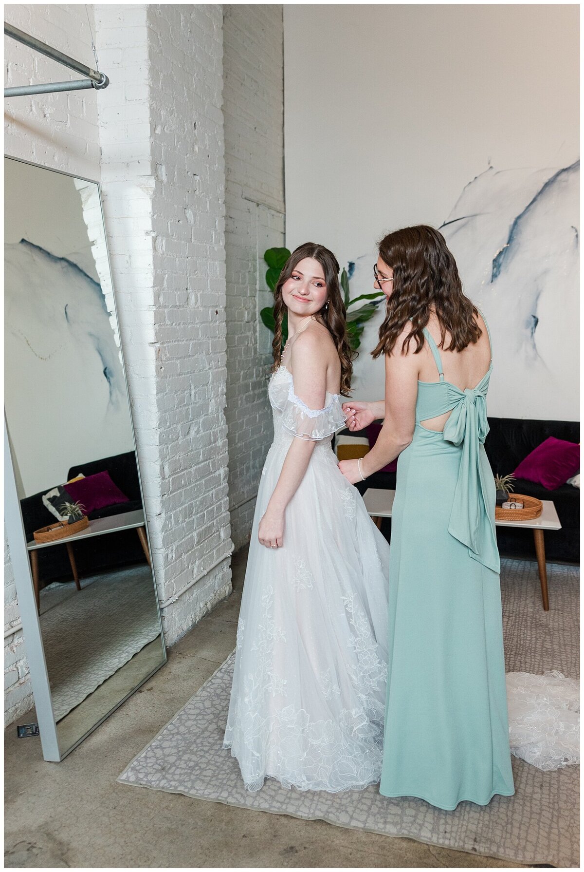 in the bridal suite of Skylight venue a bridesmaid in a light green dress helps her sister zip up the back of her wedding dress while standing in front of a mirror at Skylight Venue in Denver, captured by Denver Wedding photographer