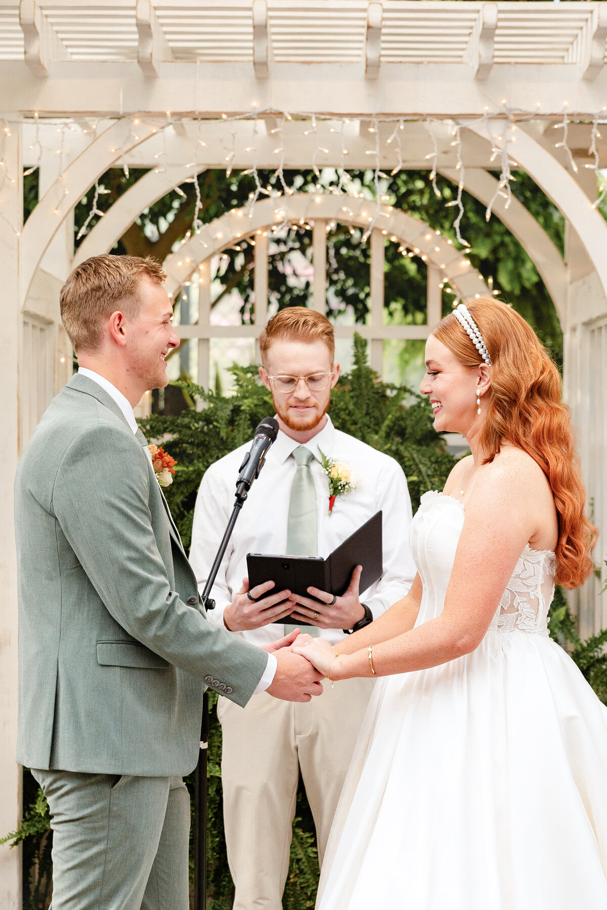 The bride and groom hold hands and exchange vows under a white wooden arch decorated with fairy lights. The officiant, wearing a matching green tie, stands behind them with a book.