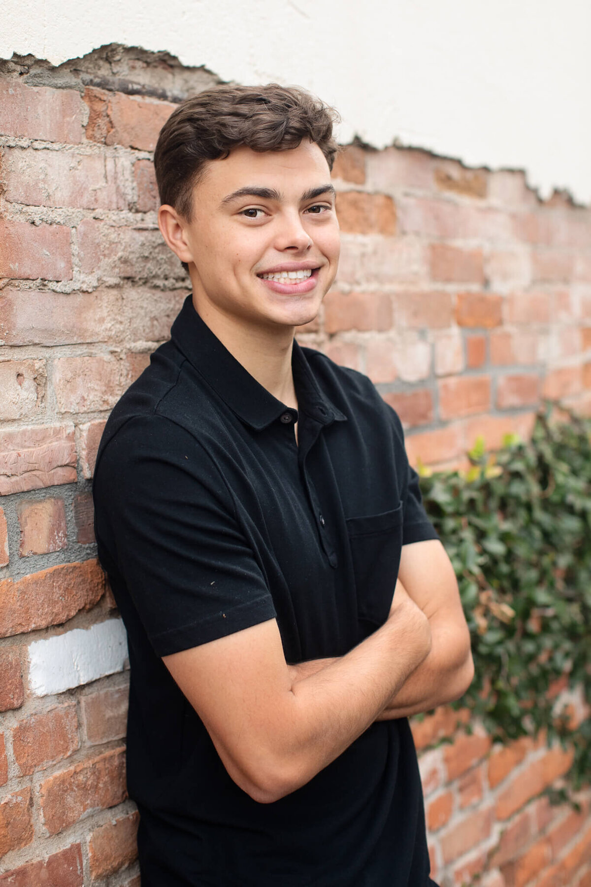 A young man in a black polo shirt leans against a brick wall, smiling with his arms crossed. Greenery is visible on the wall behind him.