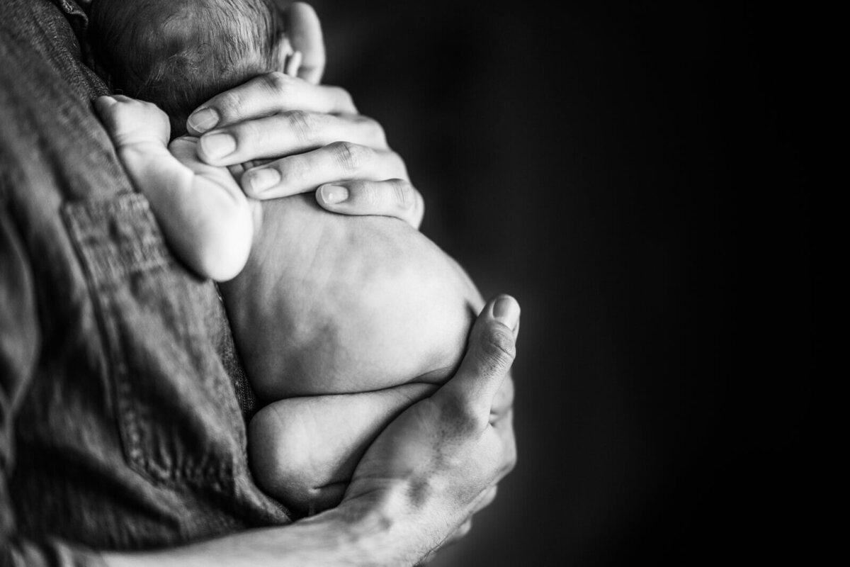 A black and white image of a newborn without a diaper cradled against his father's chest during an in home newborn photoshoot.
