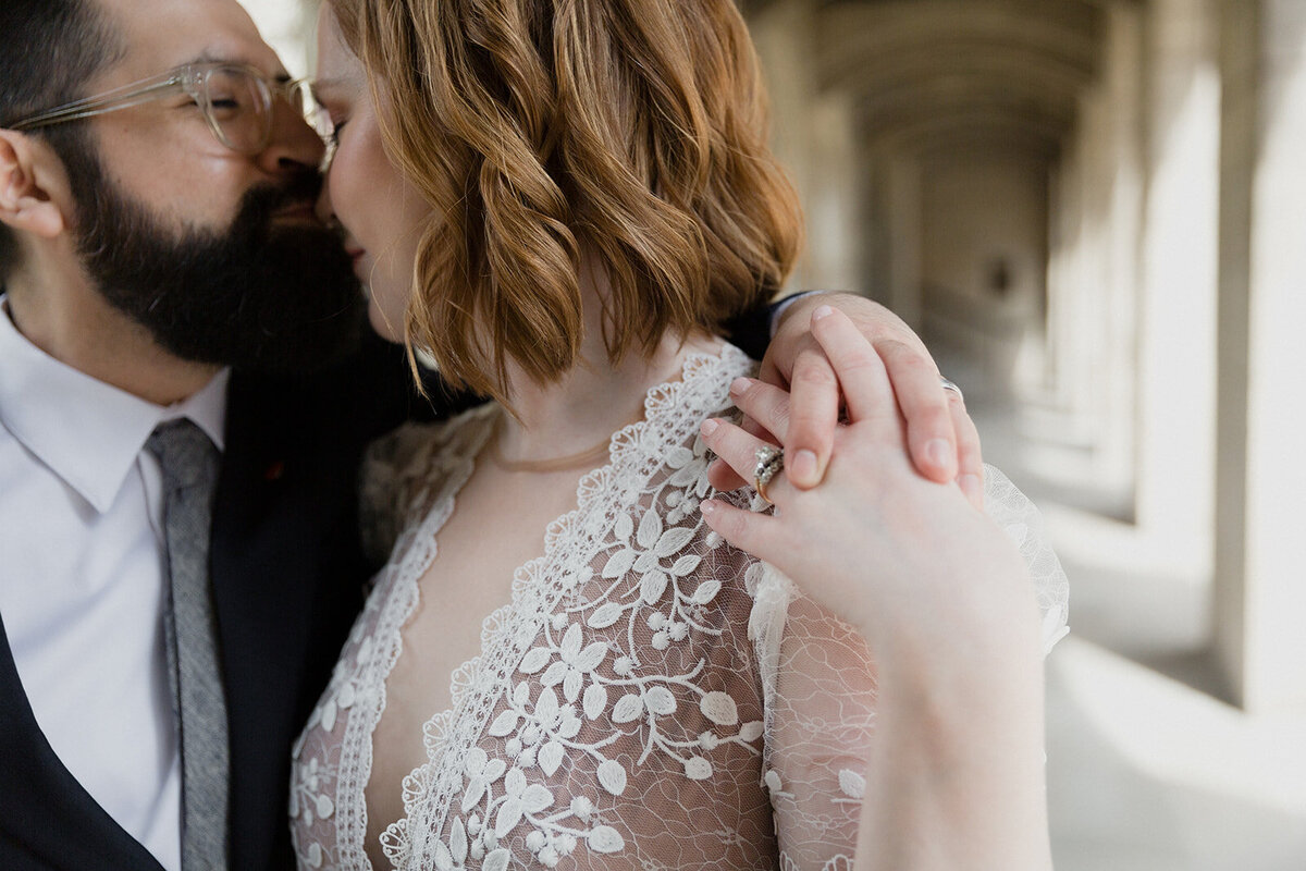 Very close up shot of wedding couple, groom's arm is over bride's shoulder and she holds his fingers, focus is on their wedding rings. Groom kisses her nose.