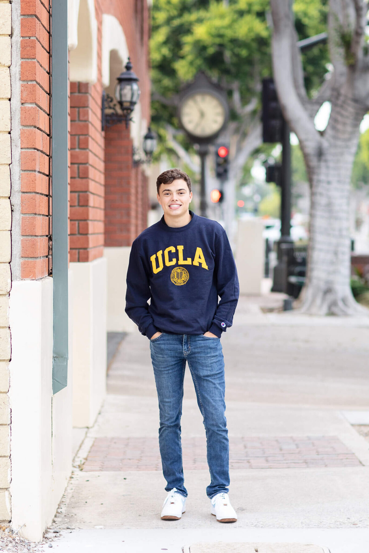 A young man stands on a sidewalk wearing a navy UCLA sweatshirt and jeans. He is smiling with hands in pockets. Theres a brick building and a vintage-style street clock in the background.