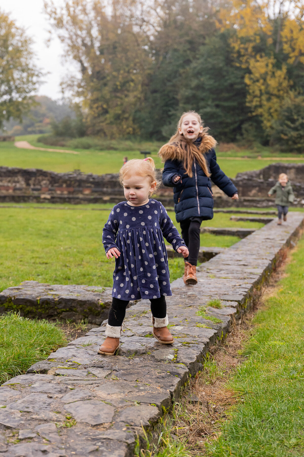 A young girl in a polka dot dress cautiously walks on a stone path, while another girl in a black jacket leaps joyfully behind her in a grassy area with autumn trees.