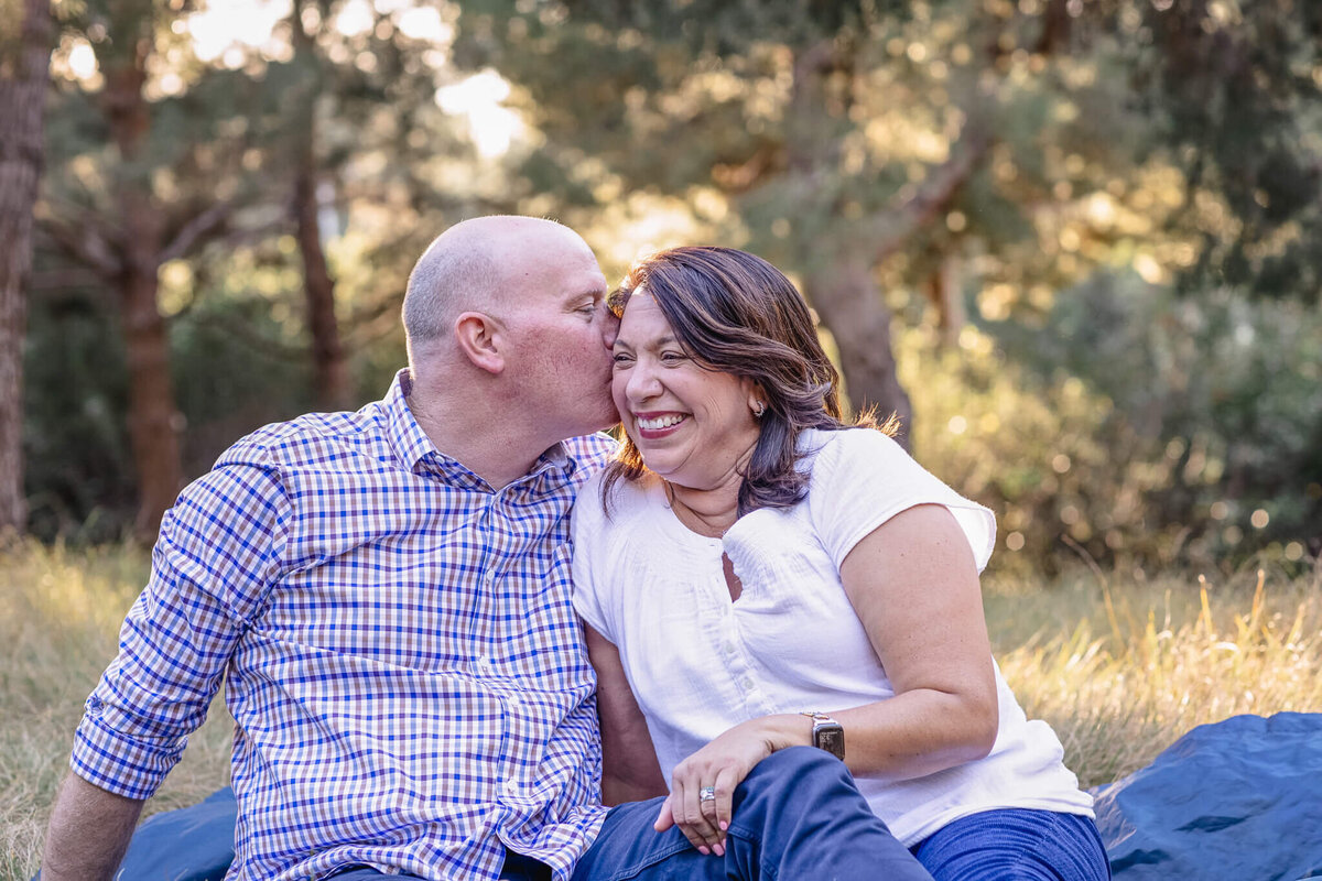 A bald man wearing a purple dress shirt kissing his wife's head while she's smiling as they sit on a navy picnic blanket in the grass.