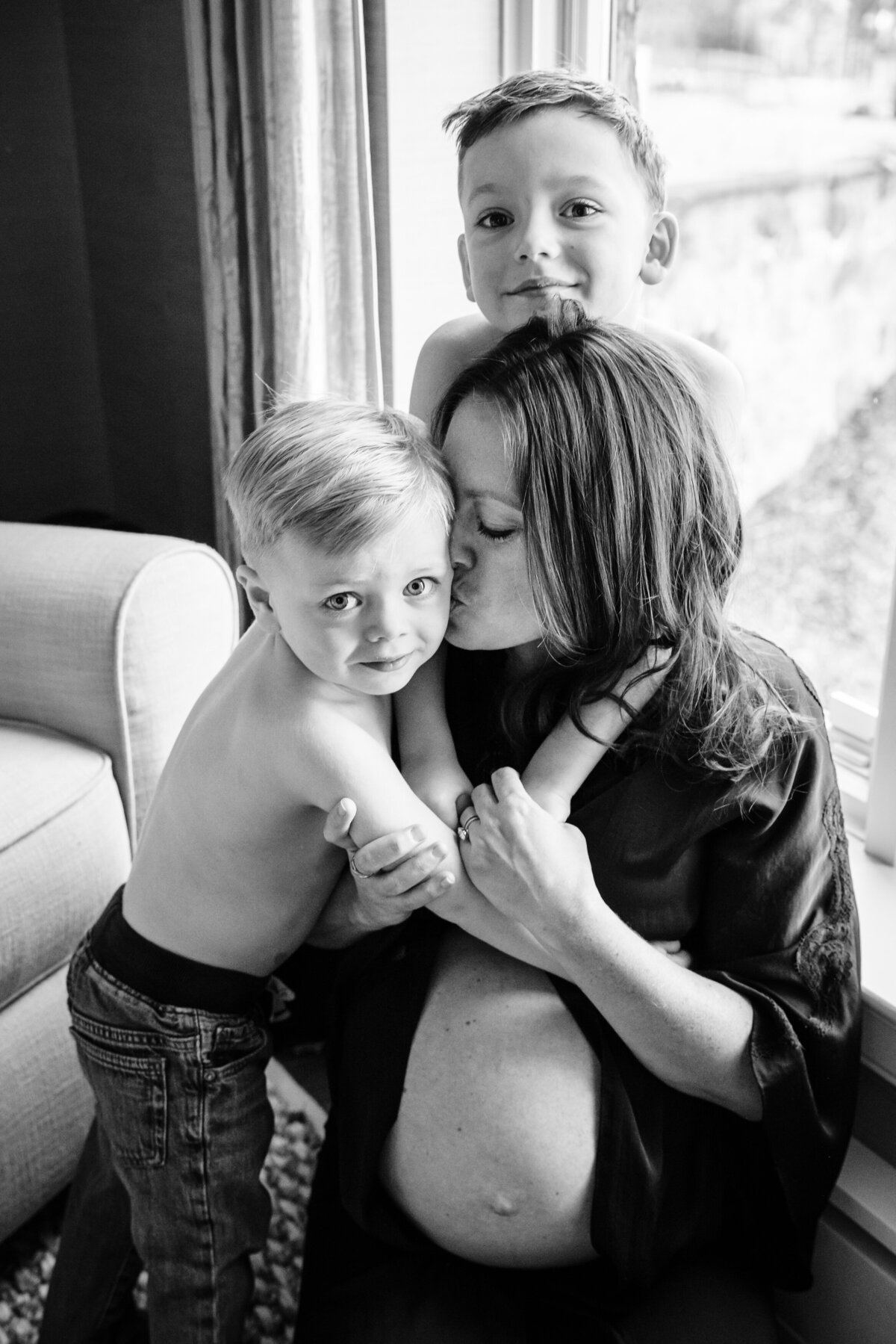 A pregnant woman in a black robe sits by a window, affectionately kissing one of her two young sons, while the other boy stands behind her, smiling.