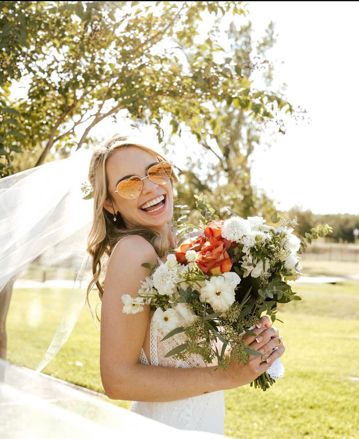 Laughing bride in heart-shaped sunglasses holding a vibrant bouquet with orange and white flowers. Her long veil flows in the breeze under sunny skies. Mistique Makeup offers mobile bridal hair and makeup services in Austin, Dallas and San Antonio, specializing in effortless, radiant looks for warm-weather weddings.
