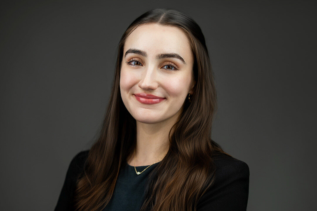 Professional headshot of a young woman with long brown hair, wearing a black blazer and dark top, smiling confidently against a gray background — photographed by Alex Kaplan Photo in New Jersey