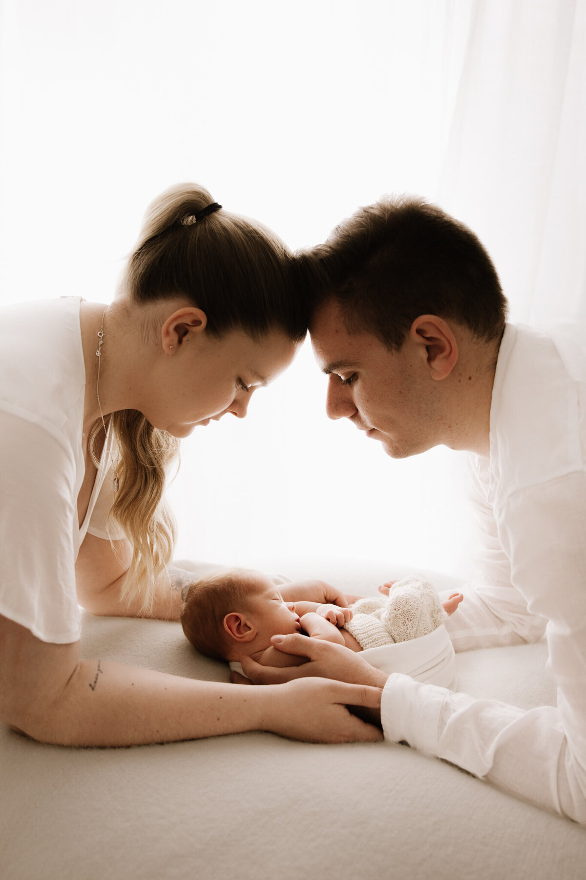 Mother and father gently resting their foreheads together as they gaze lovingly at their newborn baby lying between them. The baby is wrapped in a cream knit outfit and positioned on a soft white blanket. Captured in a Sydney photography studio with diffused artificial lighting, this image highlights connection, intimacy, and the early bond of a new family.