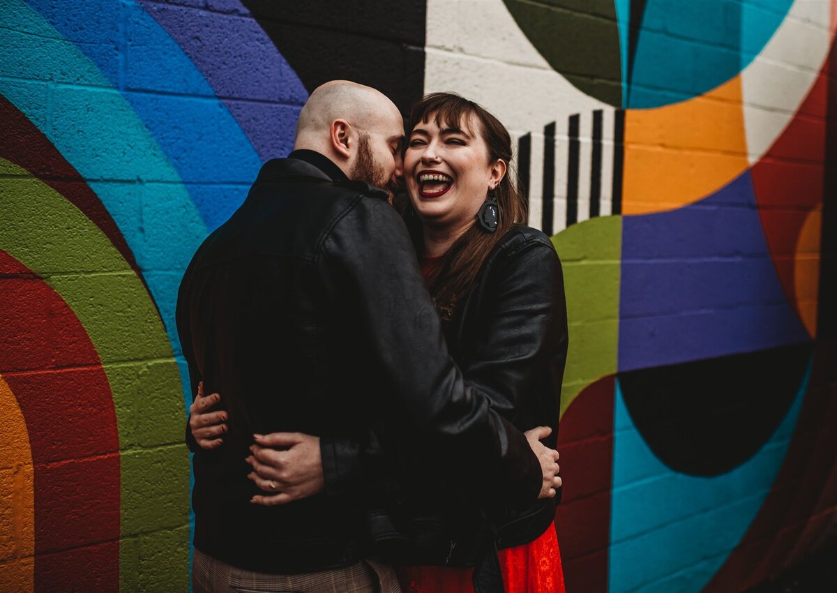 Baltimore wedding photographers captures engagement session in front of wall art in downtown with a colorful mural behind a man embracing his fiancé while she laughs