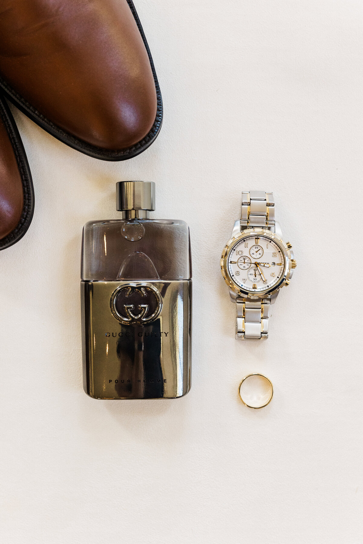 A refined flat lay of the groom's accessories, including polished brown dress shoes, a silver luxury wristwatch, wedding band, and Gucci cologne. Captured in Denton, Texas, this elegant groom’s detail photo highlights timeless style for a classic Southern wedding.