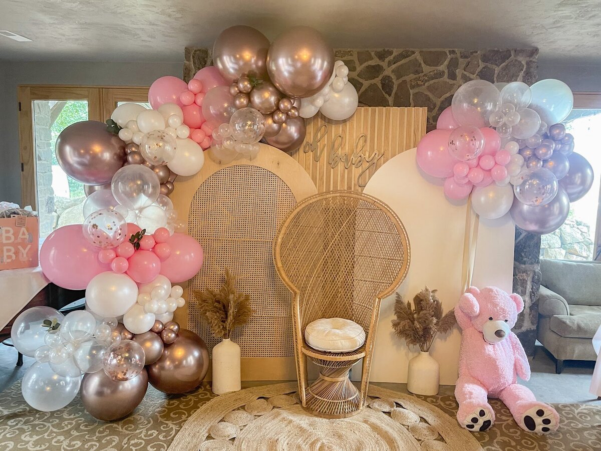 Pink, gold, and white balloons set up on wooden arch backdrops. A peacock chair and jute rug set up in front with a giant pink teddy bear.