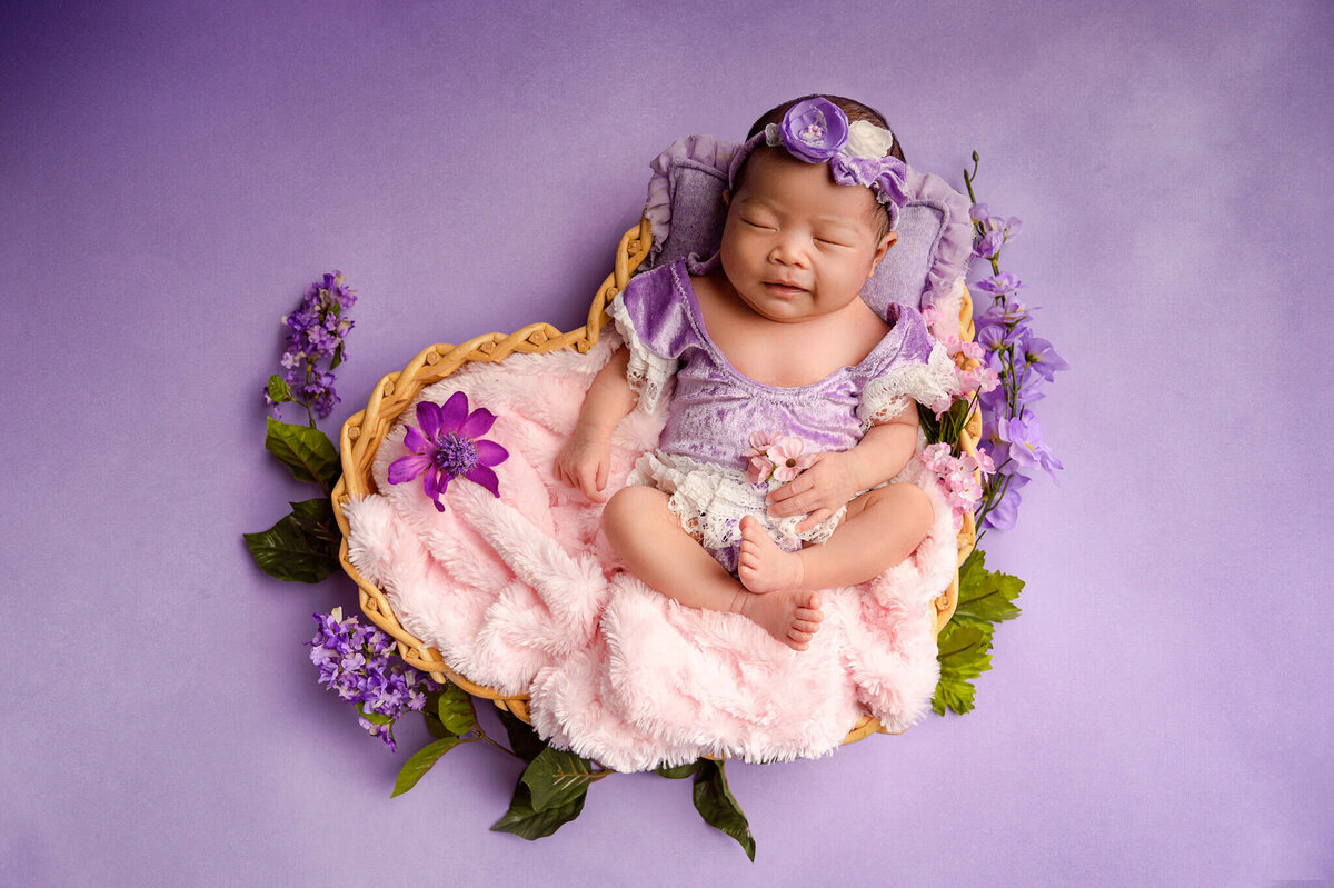 Smiling baby girl at her Greater Toronto Newborn Photo shoot in a heart basket on a purple background and purple outfit.